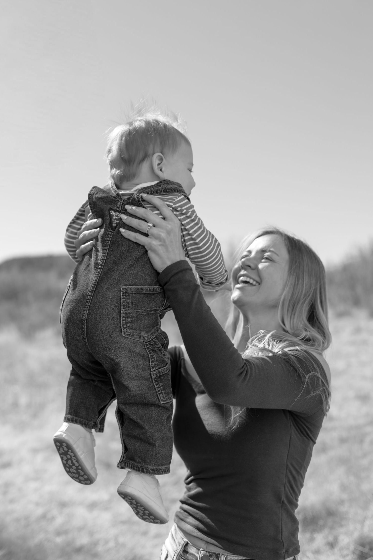 Black and white photo of a laughing mother lifting her baby in overalls outdoors on a sunny day.