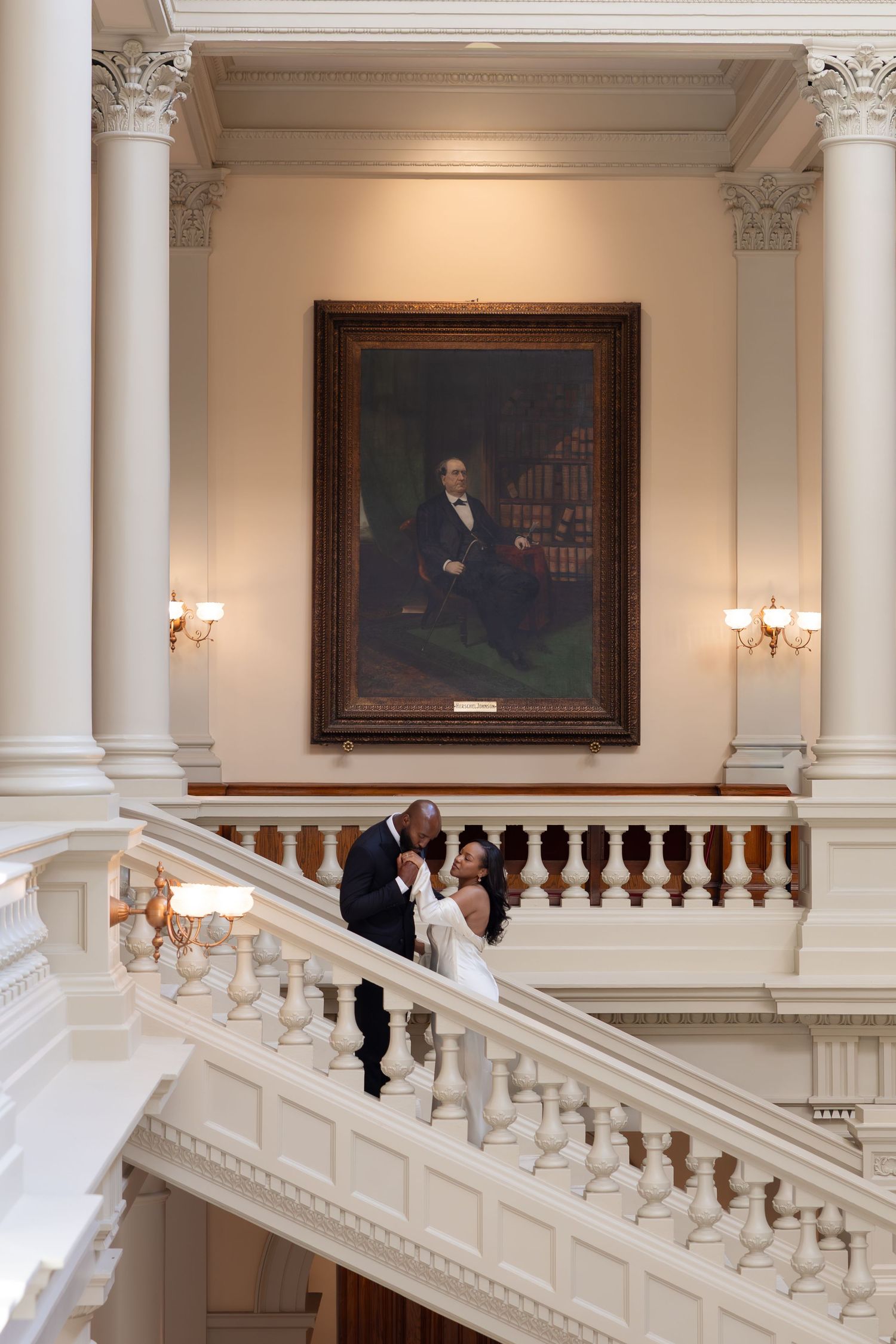 Bride and groom share a romantic moment on grand white staircase beneath a classic portrait in an elegant venue.