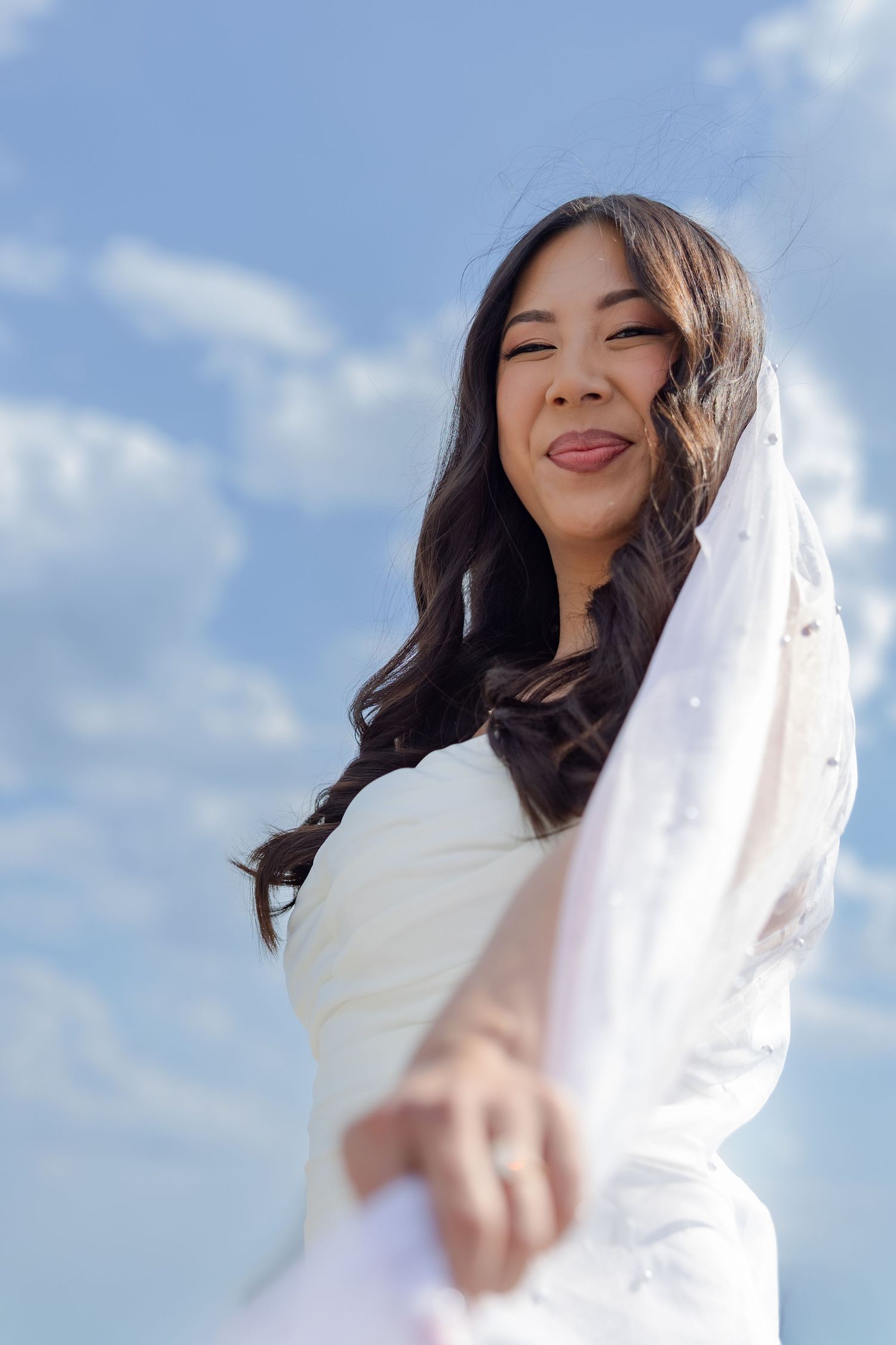Joyful Asian bride in white wedding dress and veil smiling against blue sky.