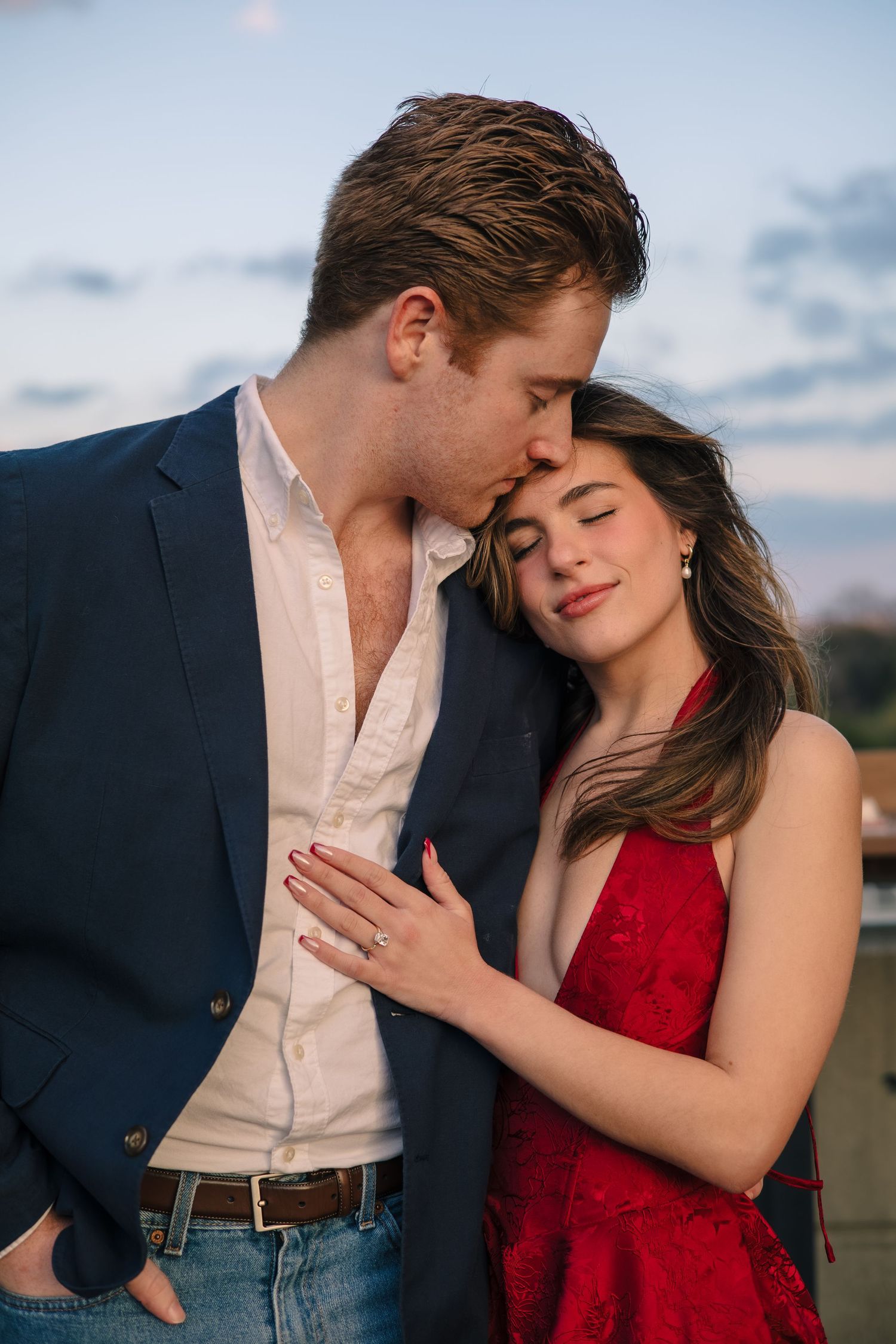 Intimate engagement portrait of couple in formal wear against sunset sky.