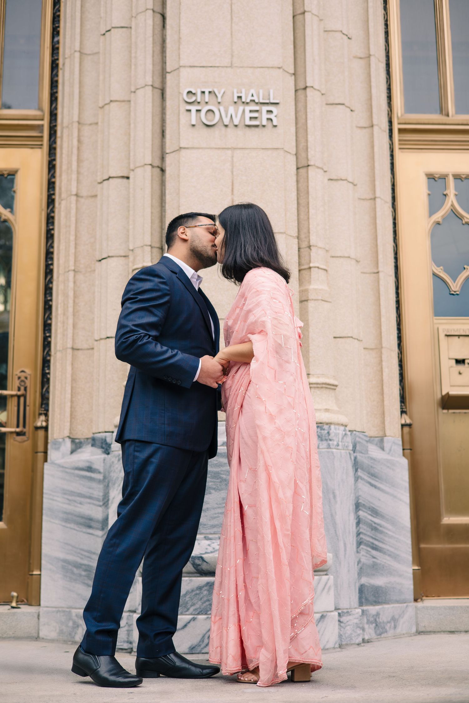 A couple shares a kiss in front of City Hall Tower wearing formal attire.