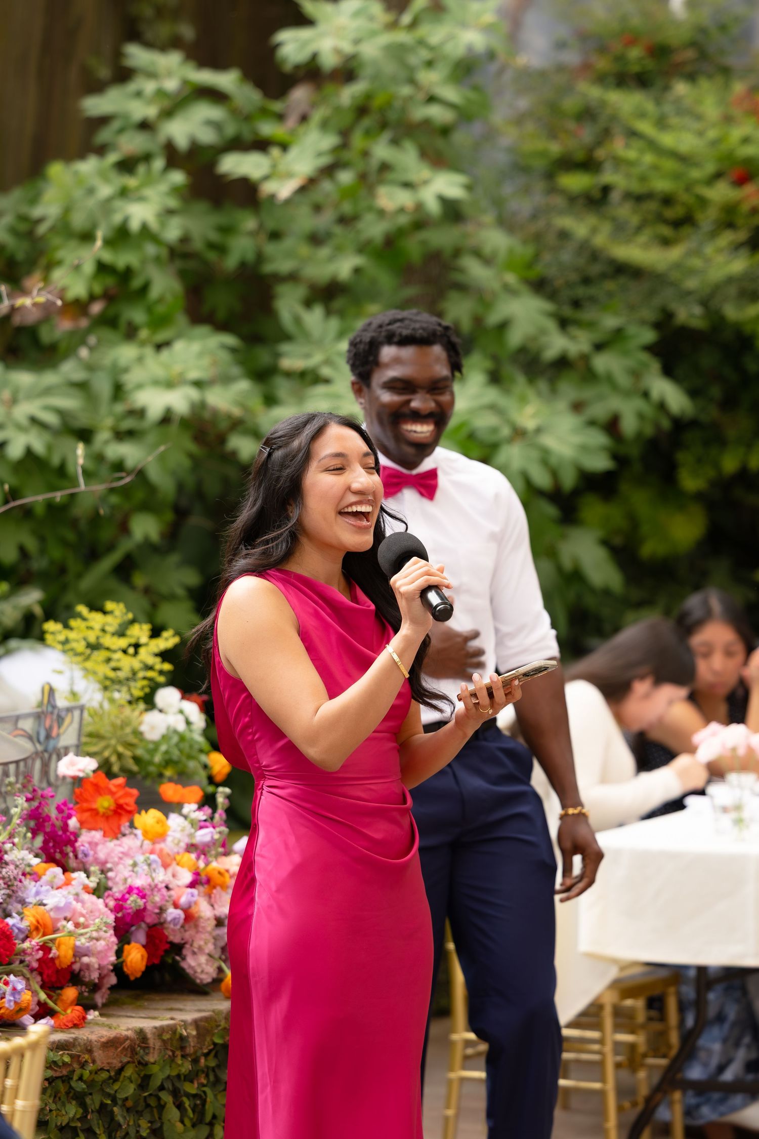 Woman in pink dress giving speech at outdoor wedding reception with man in bow tie smiling behind her.