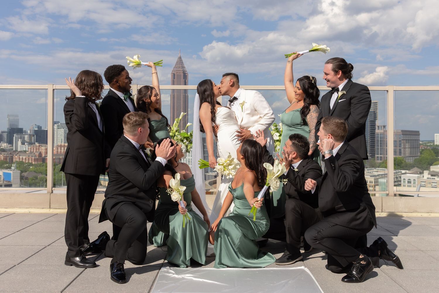 Wedding party surrounds kissing couple on rooftop with city skyline, bridesmaids in sage green gowns.