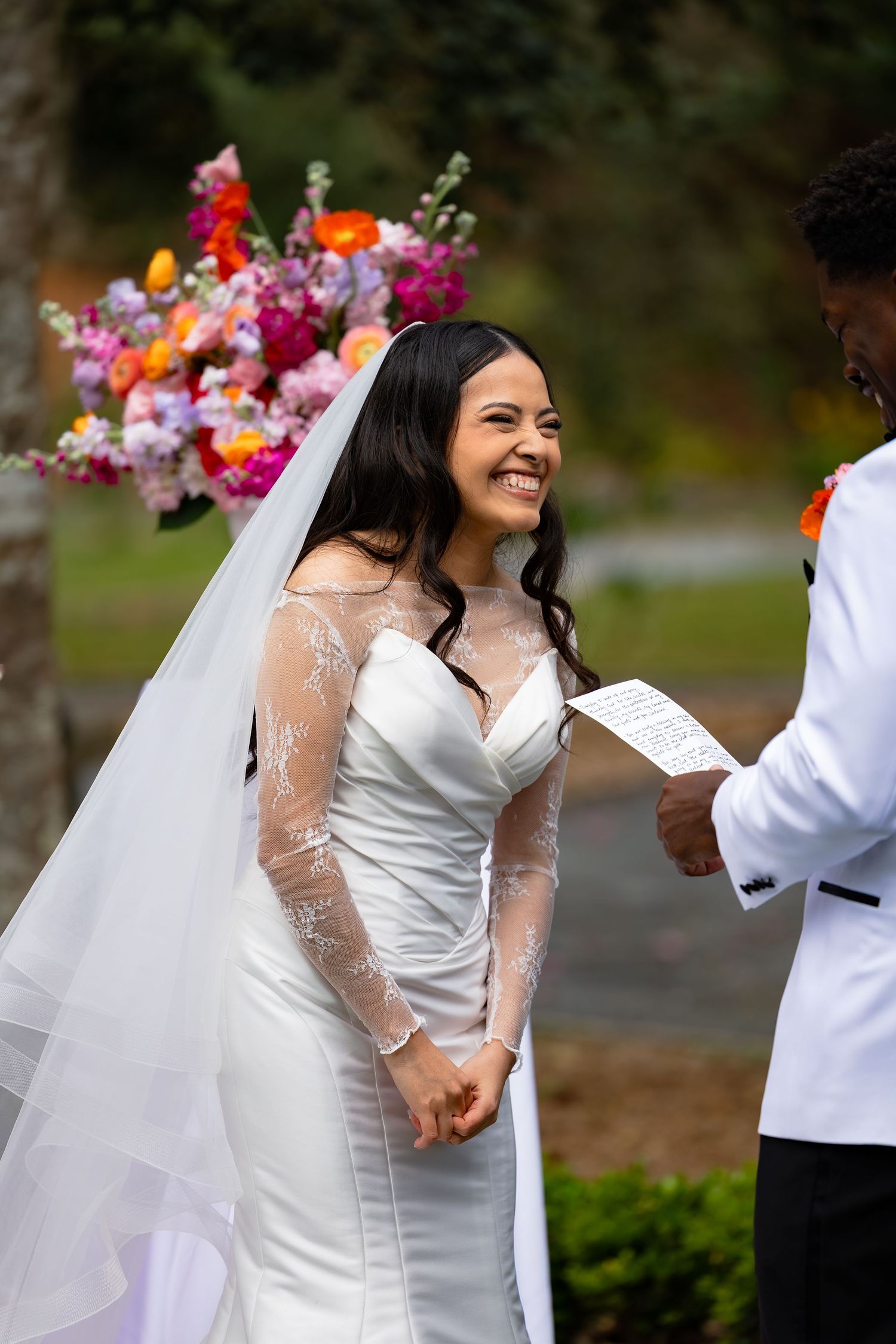 Smiling bride in white lace gown and veil holds hands with groom in white suit during outdoor wedding ceremony.