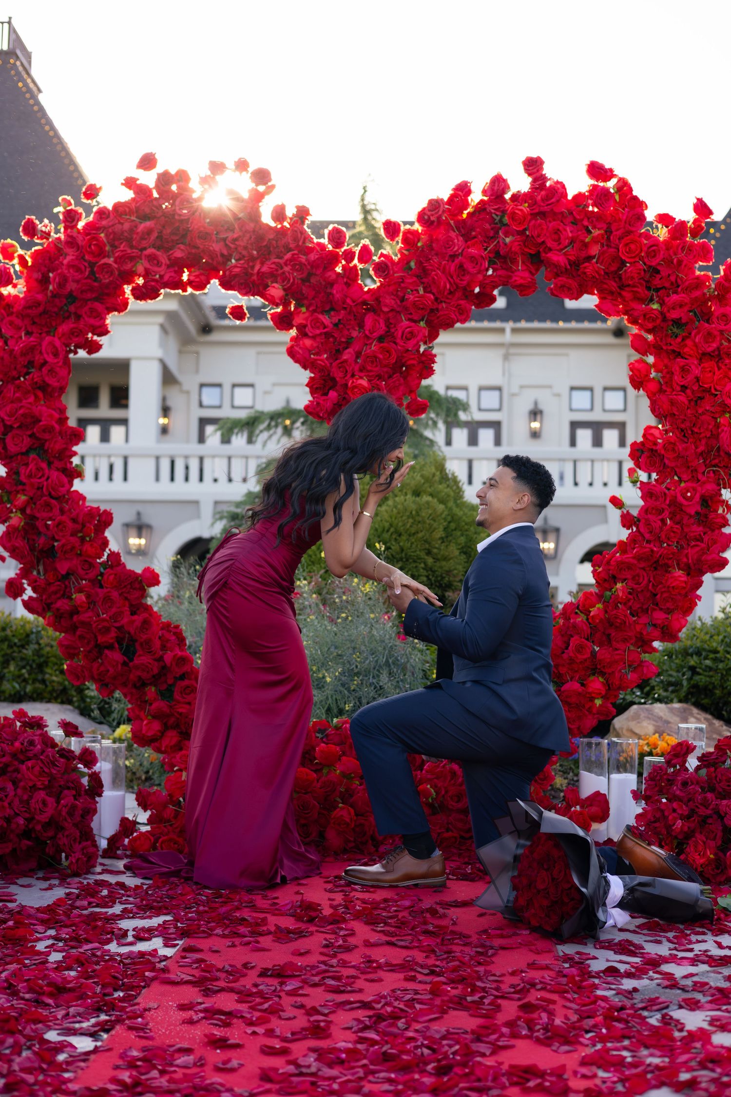 Man proposes to woman under a large red rose heart arch, with rose petals on the ground, at a romantic outdoor setting.
