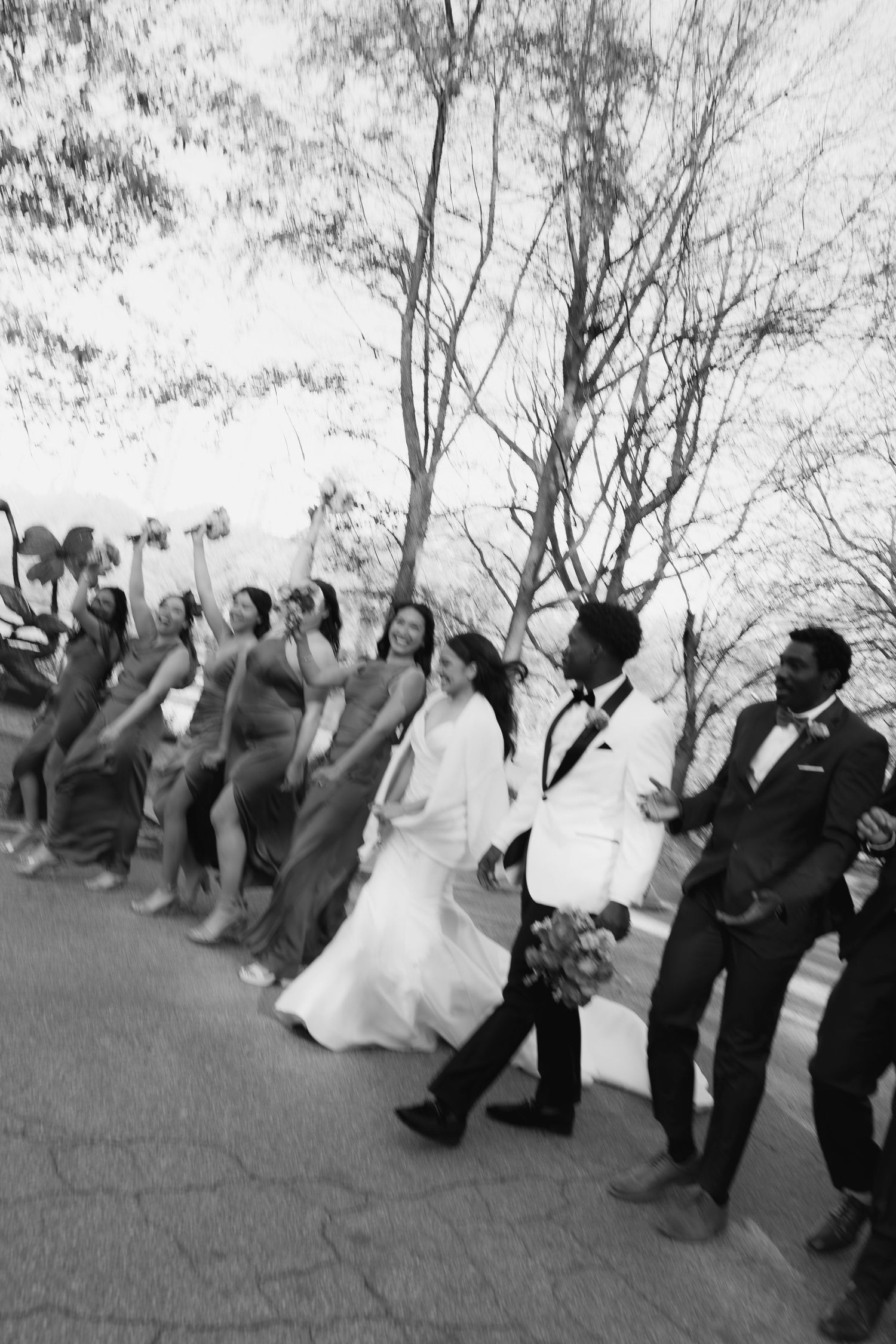 Wedding party celebrates in black and white photo, bridesmaids in gowns and groomsmen in suits cheering outdoors.