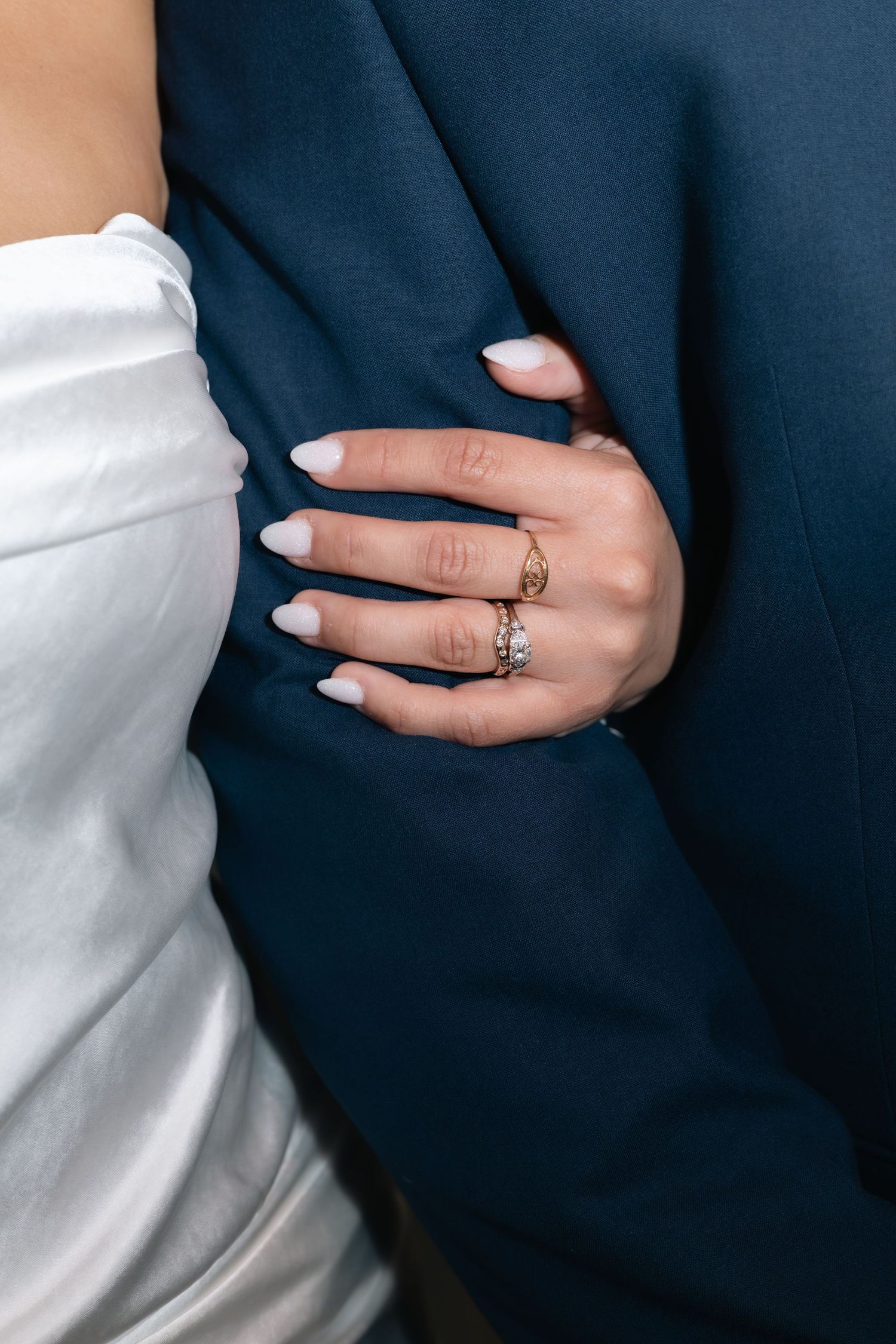 Close-up of manicured hand with wedding ring against navy suit and white dress fabric.