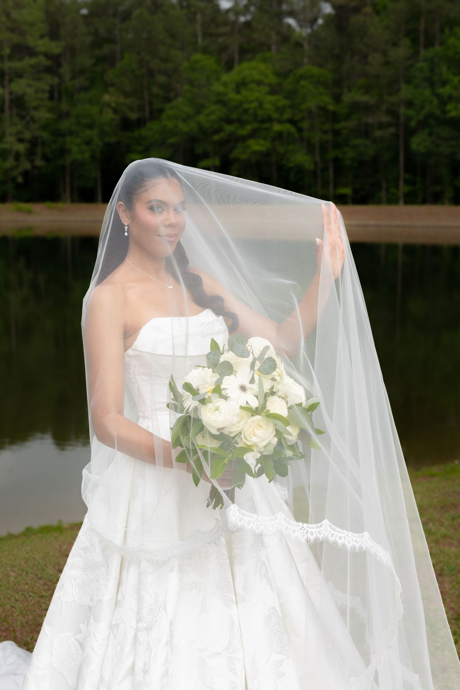 Bride in white strapless gown holds bouquet of white roses, lifting her veil near a serene lake backdrop.