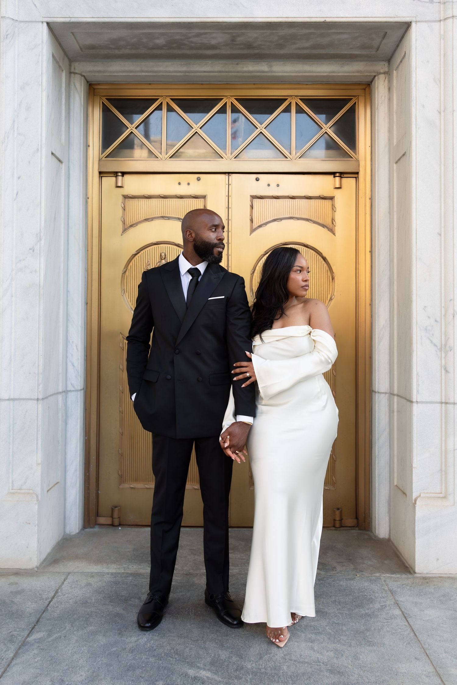 Elegant couple posing before golden doors, man in black suit, woman in white off-shoulder gown, formal engagement photo.