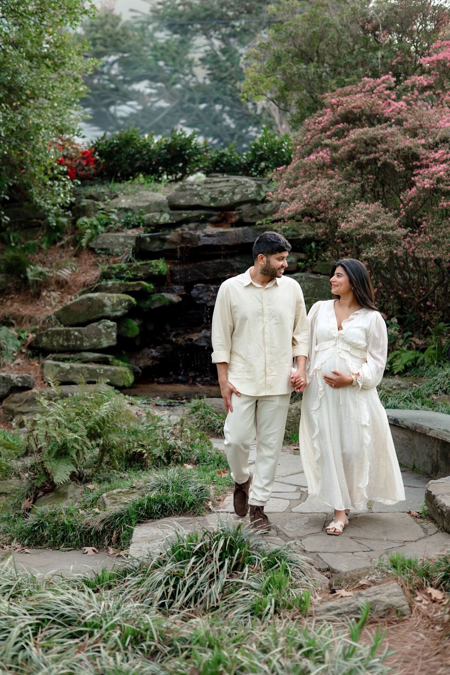 Couple in matching cream outfits share romantic moments in a lush garden setting with blooming flowers and stone features.
