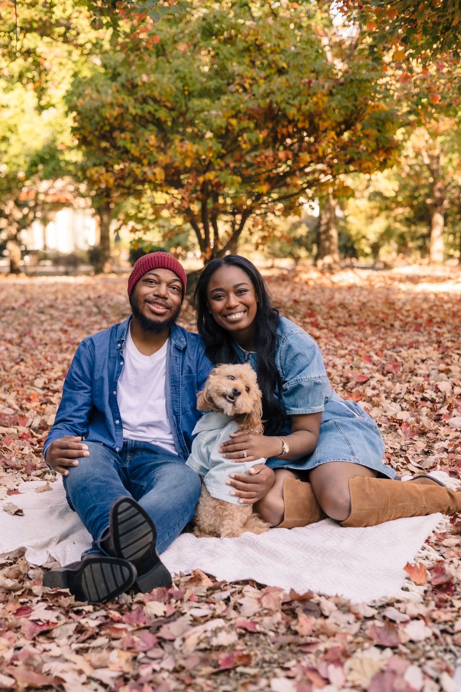 A couple relaxes on a blanket with their small dog among fallen autumn leaves.