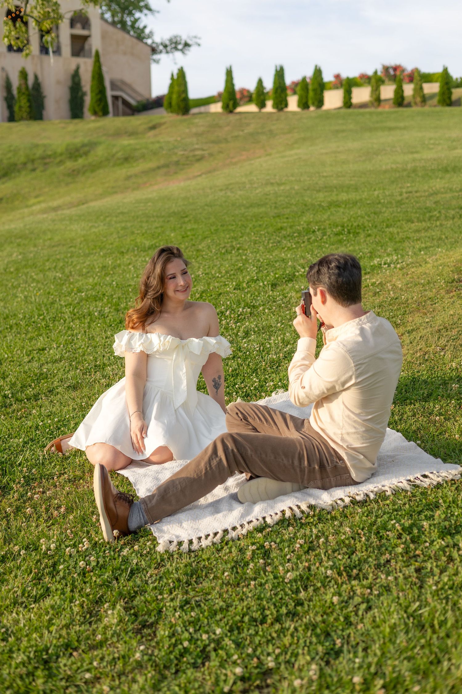 Couple enjoying a romantic picnic on green grass, woman in white dress smiling as man photographs her outdoors.