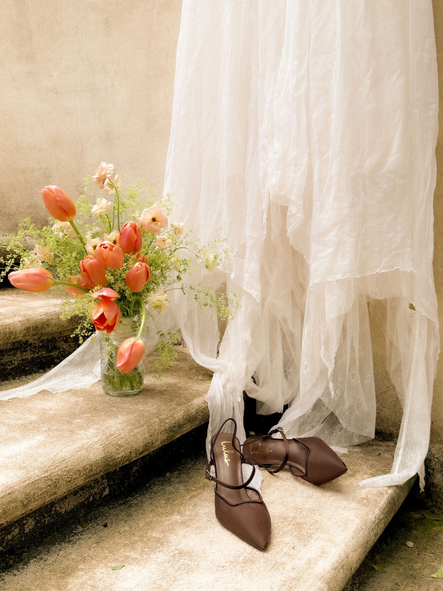 A wedding dress hem and brown bridal shoes beside coral tulips in a vintage setting.
