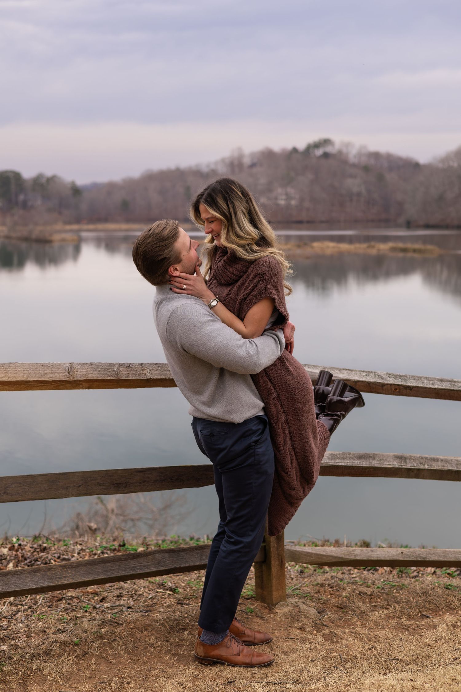 A couple shares a romantic moment along a lakeside wooden railing at sunset.