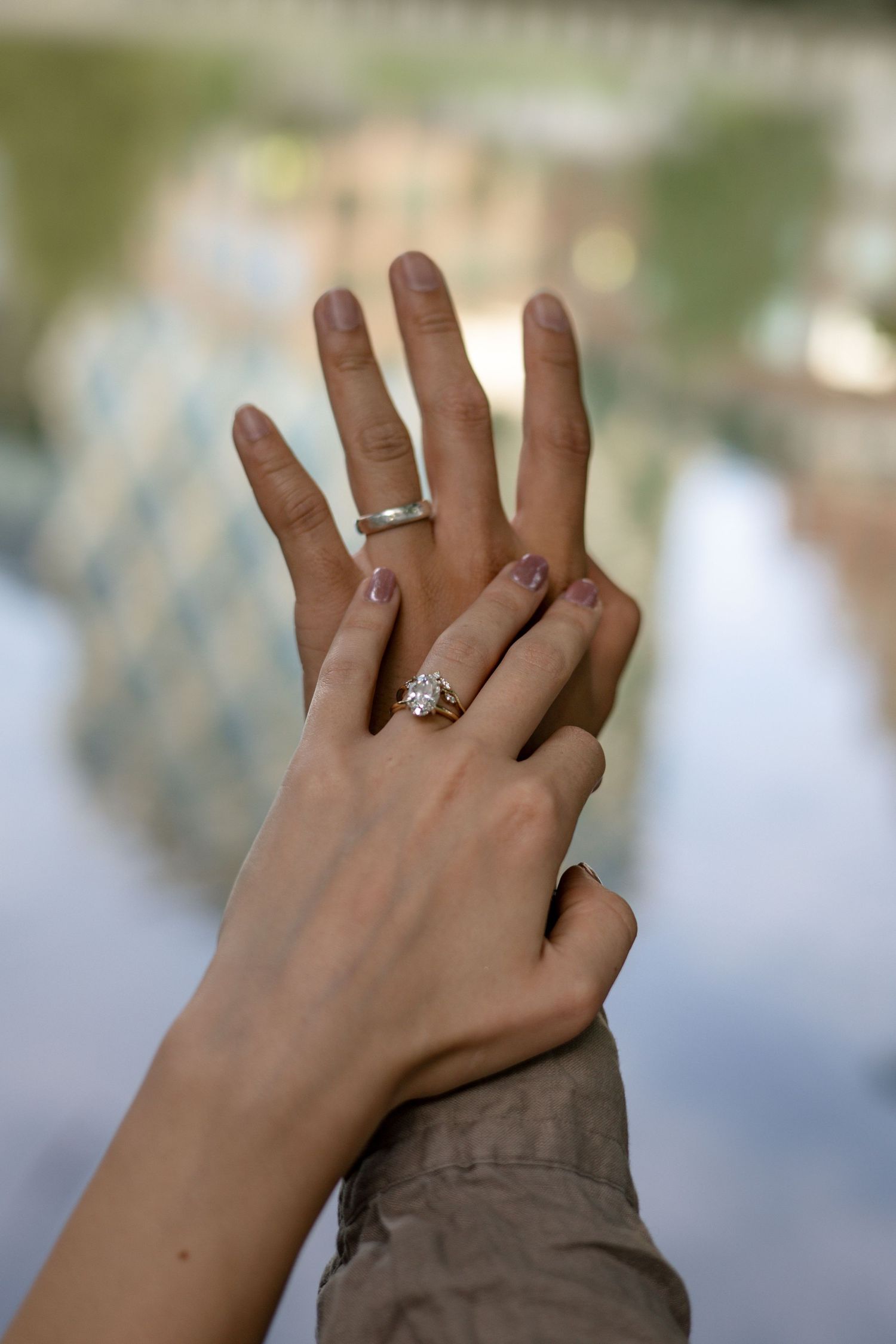 Two hands with engagement rings intertwined against a blurred nature background.