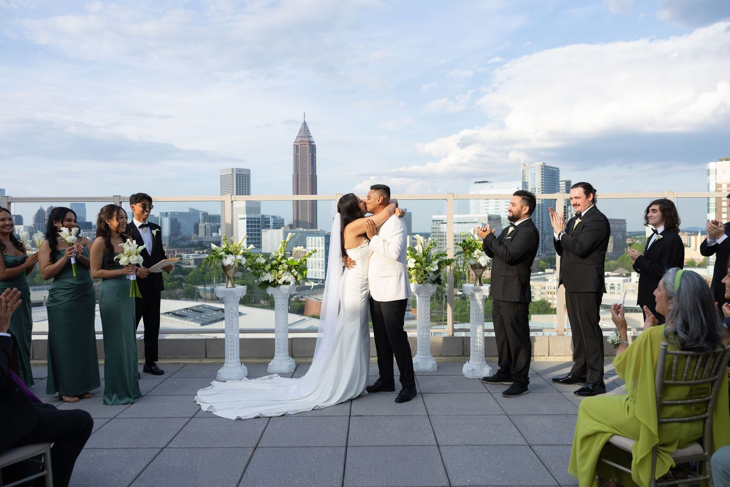 Couple shares first kiss at rooftop wedding ceremony with Atlanta skyline backdrop, surrounded by guests in elegant attire.