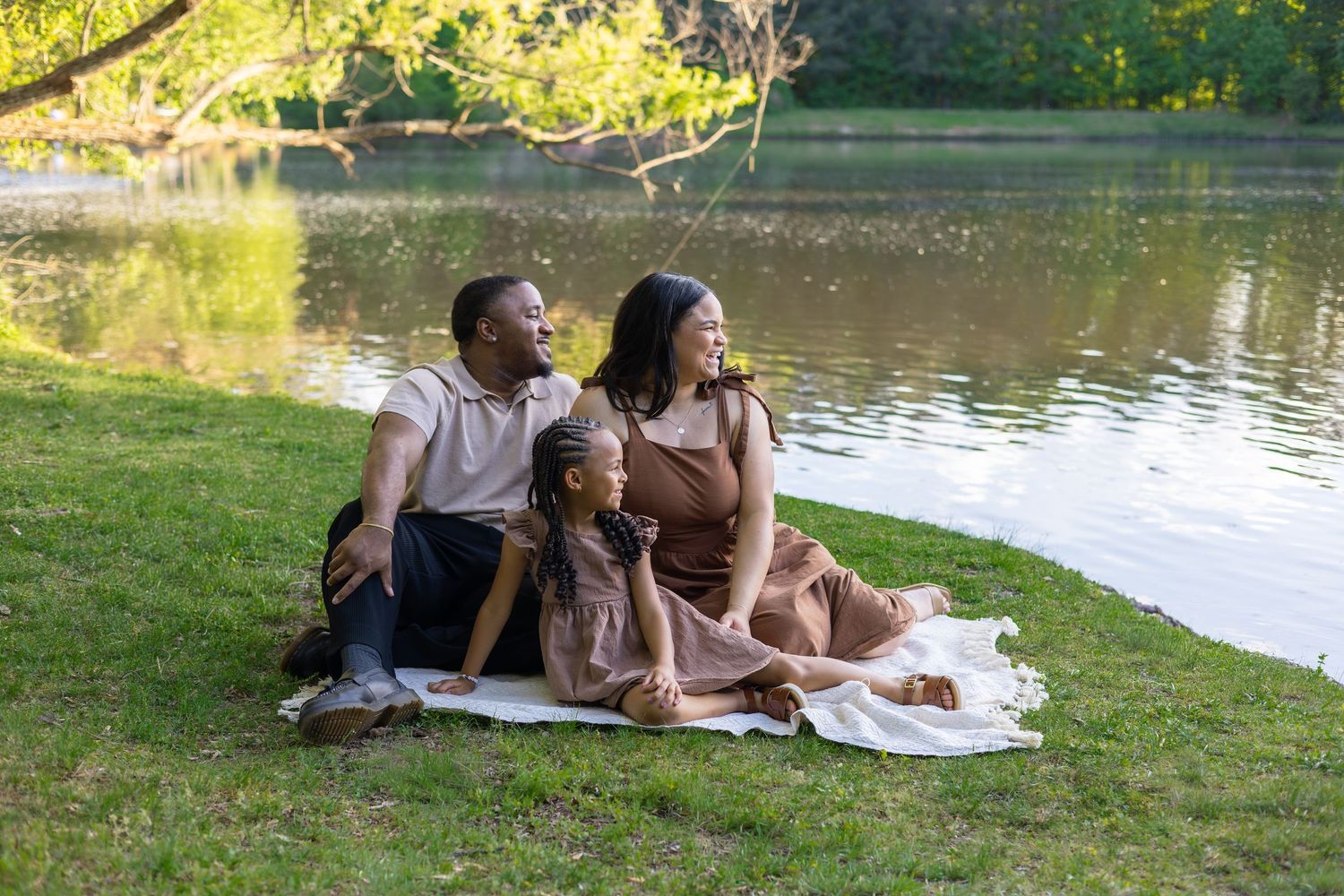A joyful family of three sits on a blanket by a serene lake, smiling together on a sunny day.