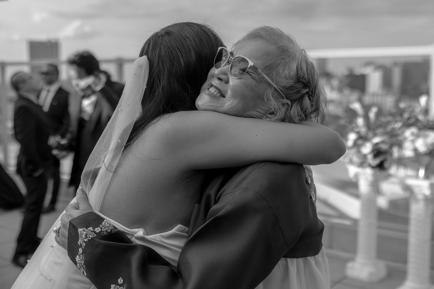Black and white photo of a bride sharing an emotional hug with a smiling wedding guest outdoors.
