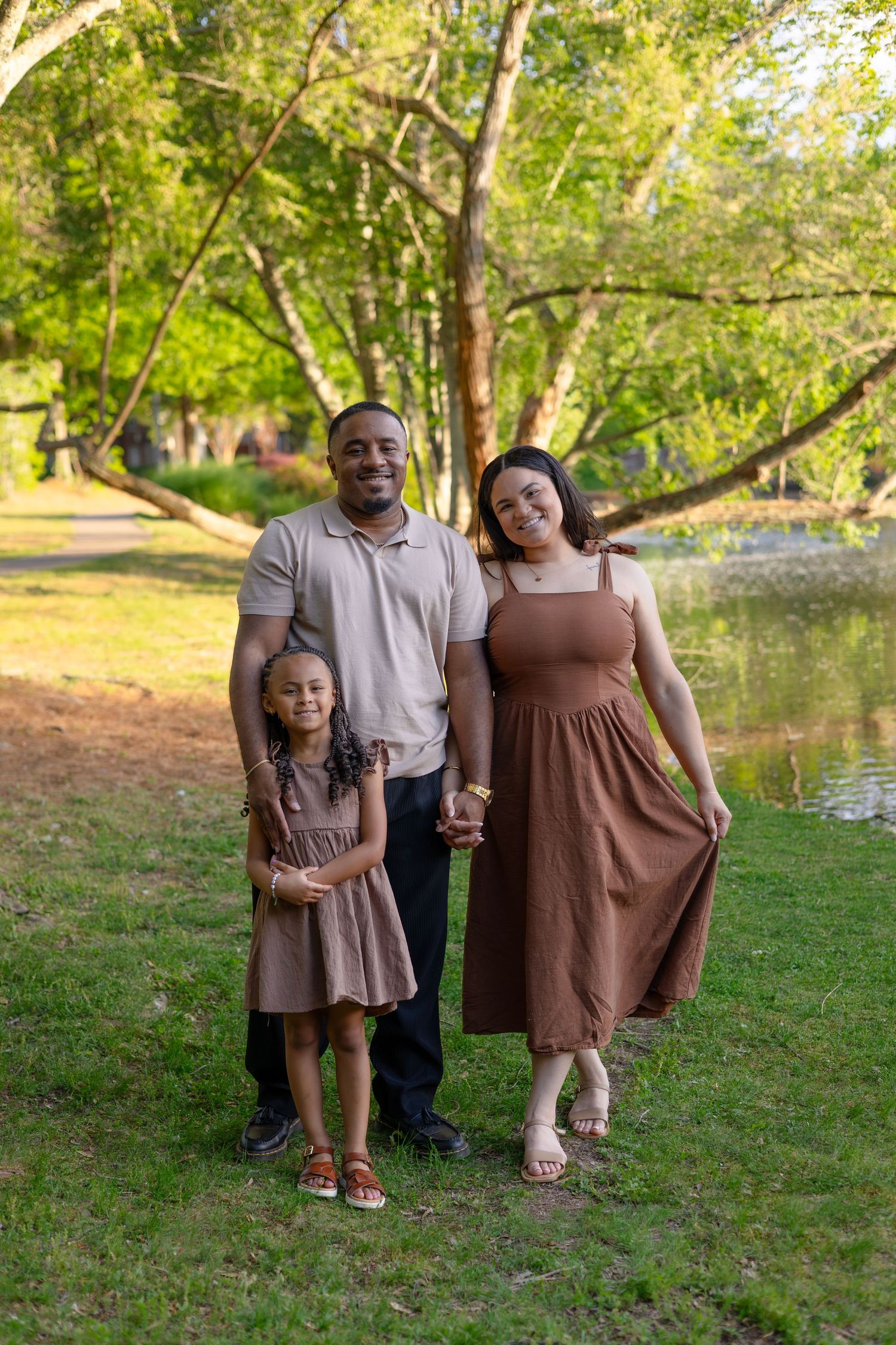 A smiling family of three poses outdoors near a pond, wearing coordinating brown and tan outfits in a lush green park.