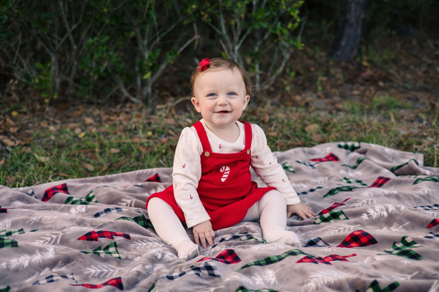 Baby in red and white outfit sitting on patterned blanket outdoors.