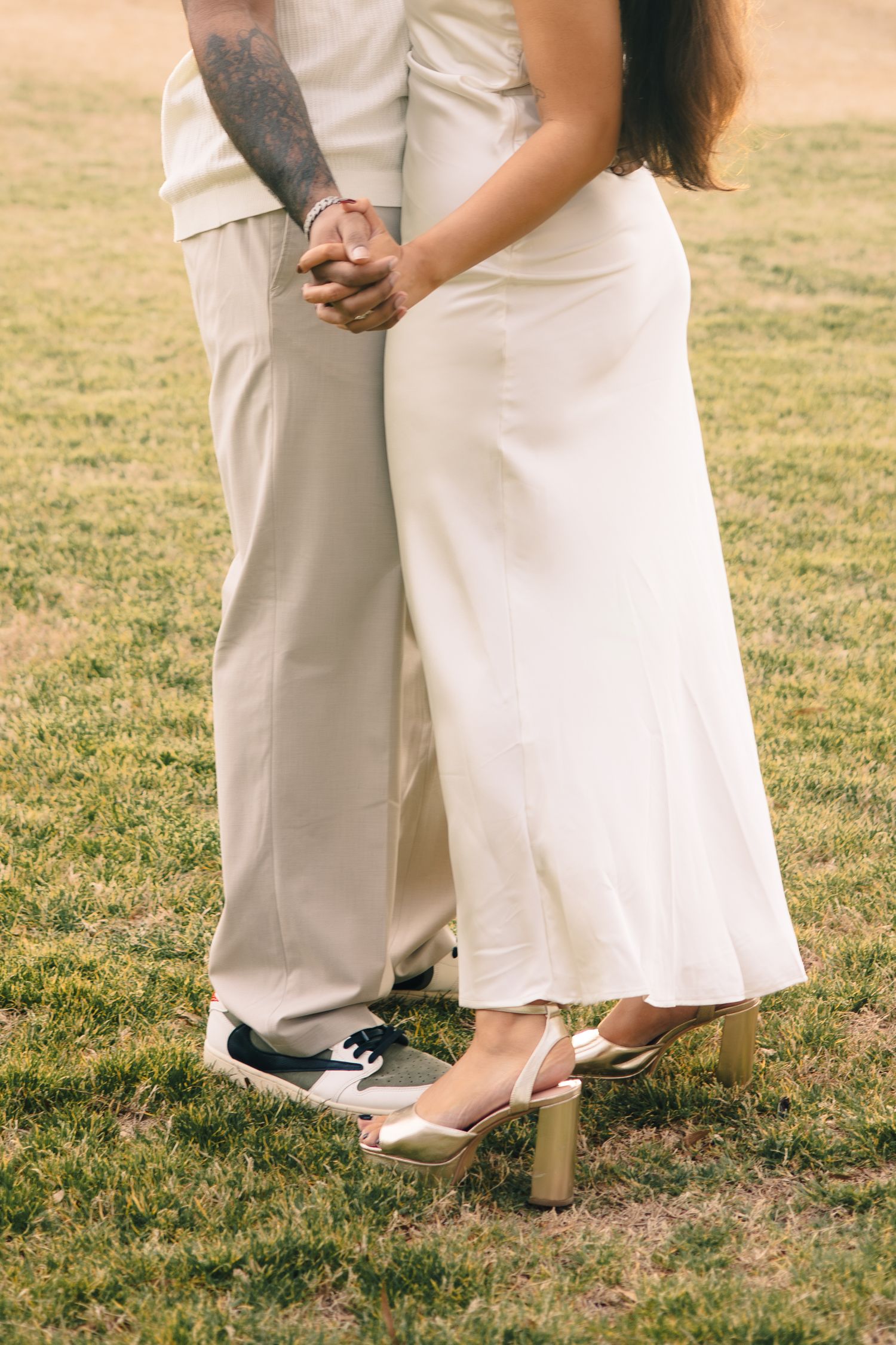 A close-up of two people in white attire holding hands while standing on grass, showing only their lower bodies and feet.