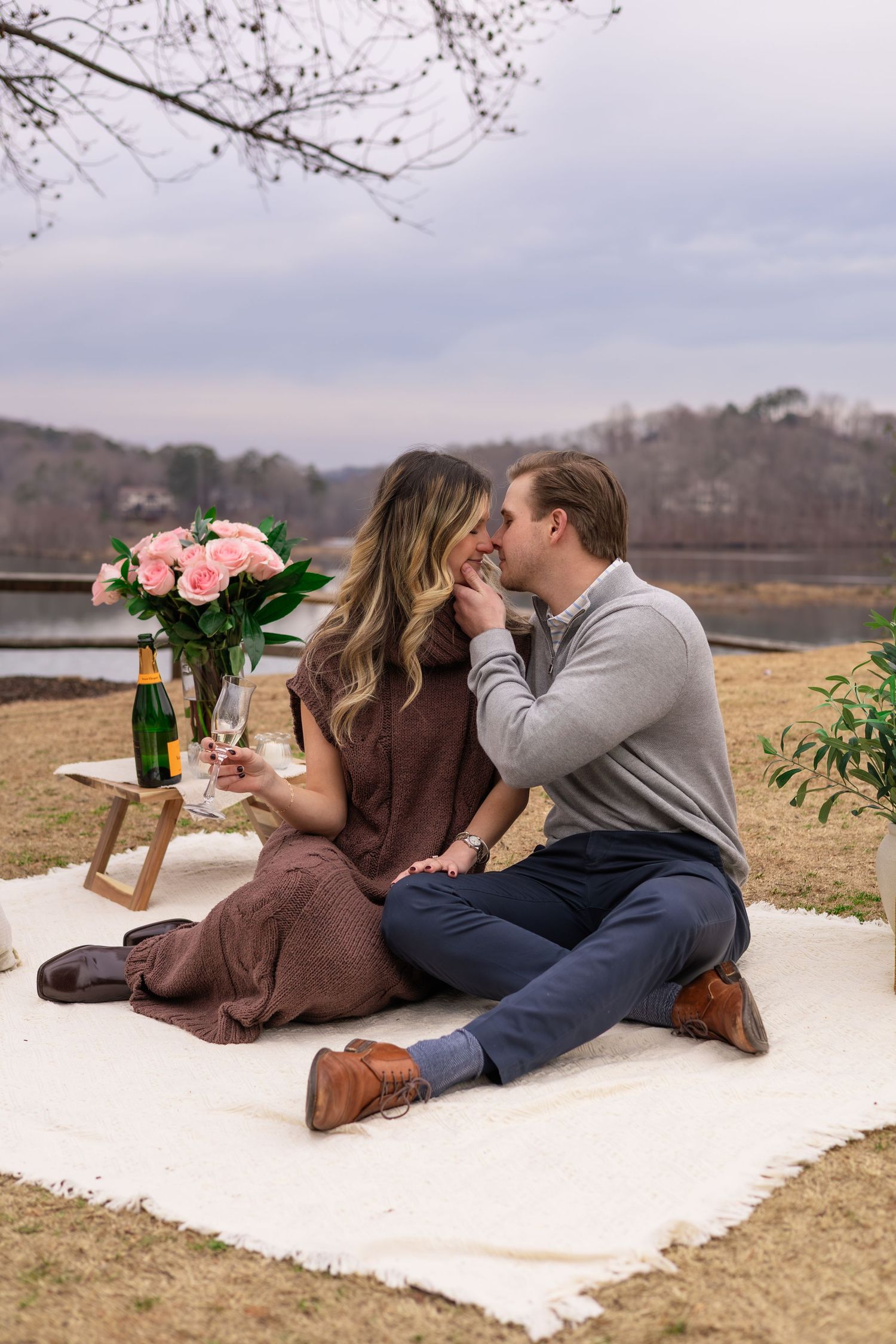A couple lounges on a white blanket with pink flowers and champagne during an outdoor picnic.