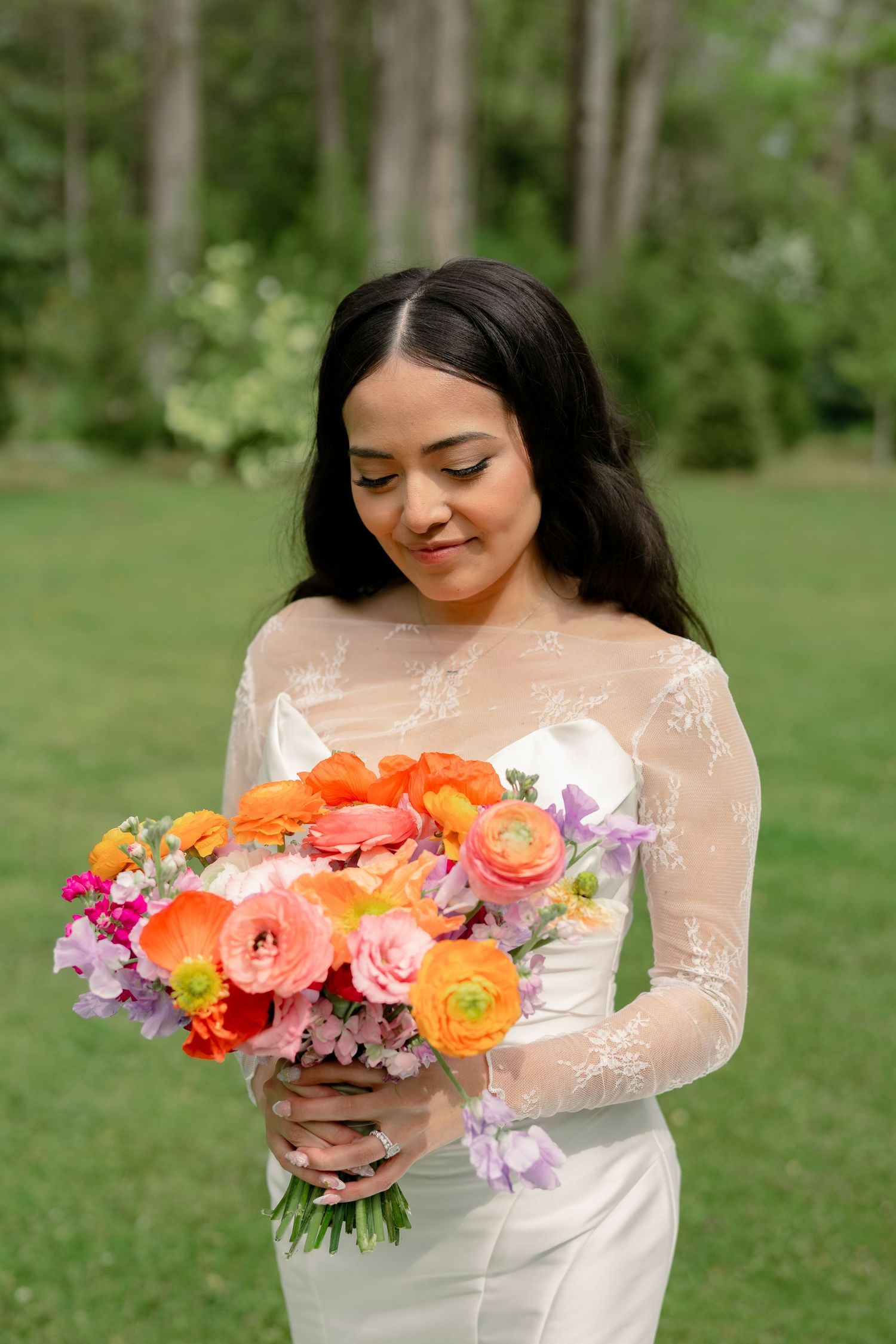 Bride in a lace-sleeved white gown holds a vibrant orange and pink floral bouquet outdoors.
