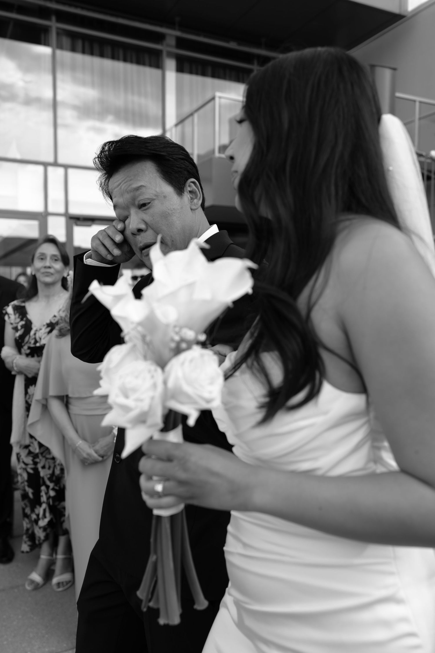 Emotional black and white photo of father wiping tears as bride holds white rose bouquet at wedding.