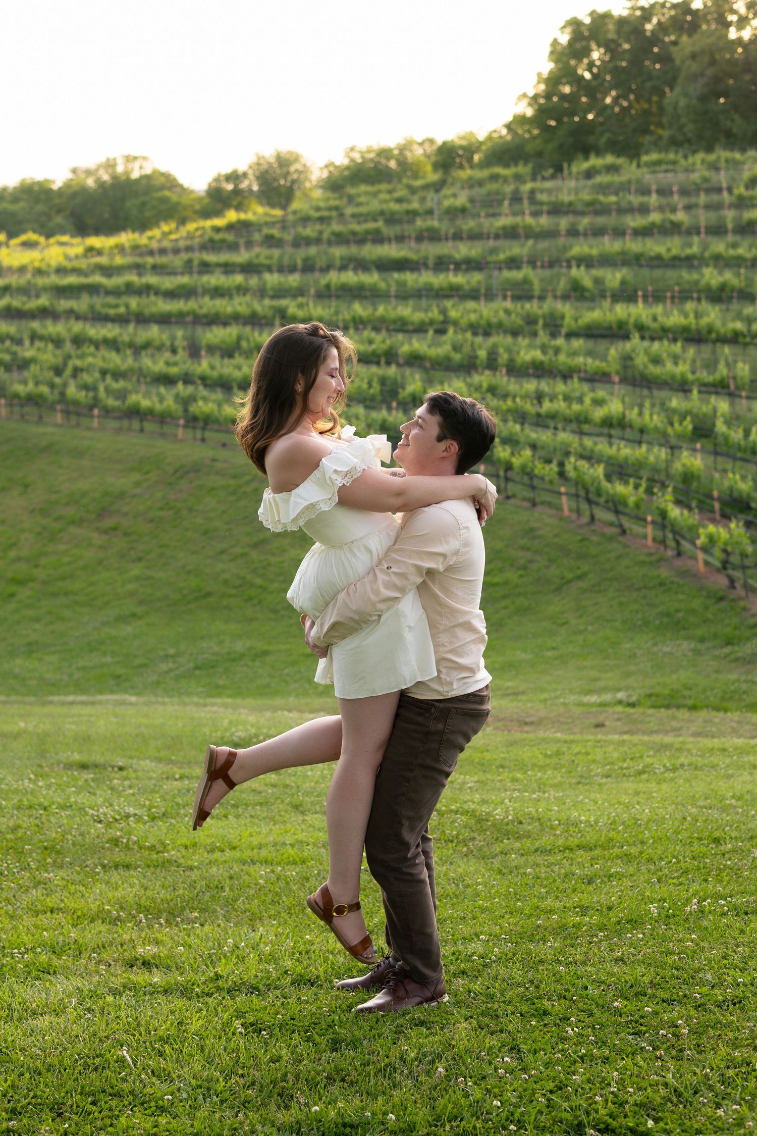 Couple embracing in a vineyard at golden hour, man lifting woman in white off-shoulder dress on green grass.