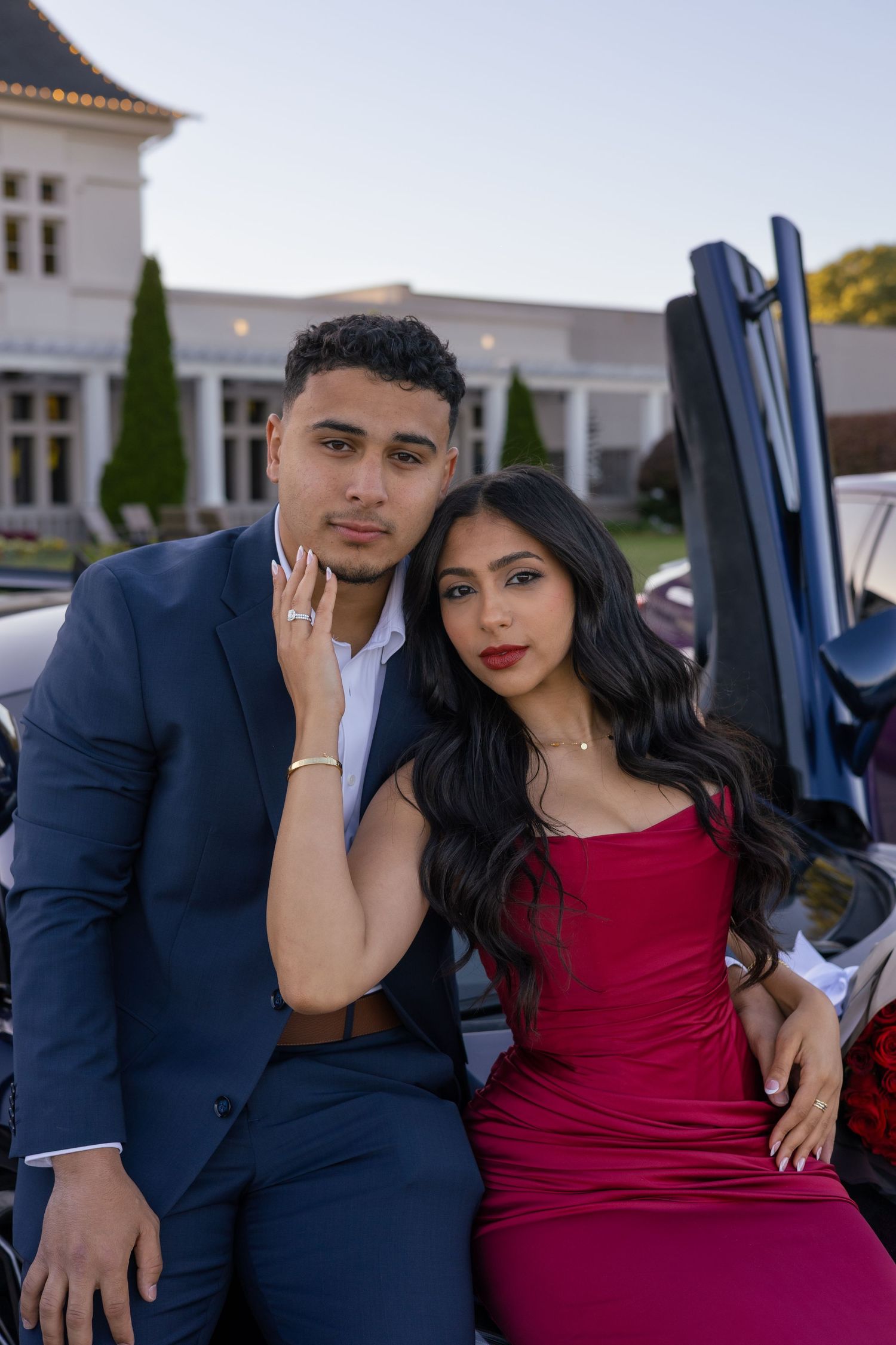 A young couple in formal attire poses by a luxury car, man in navy suit, woman in red dress.