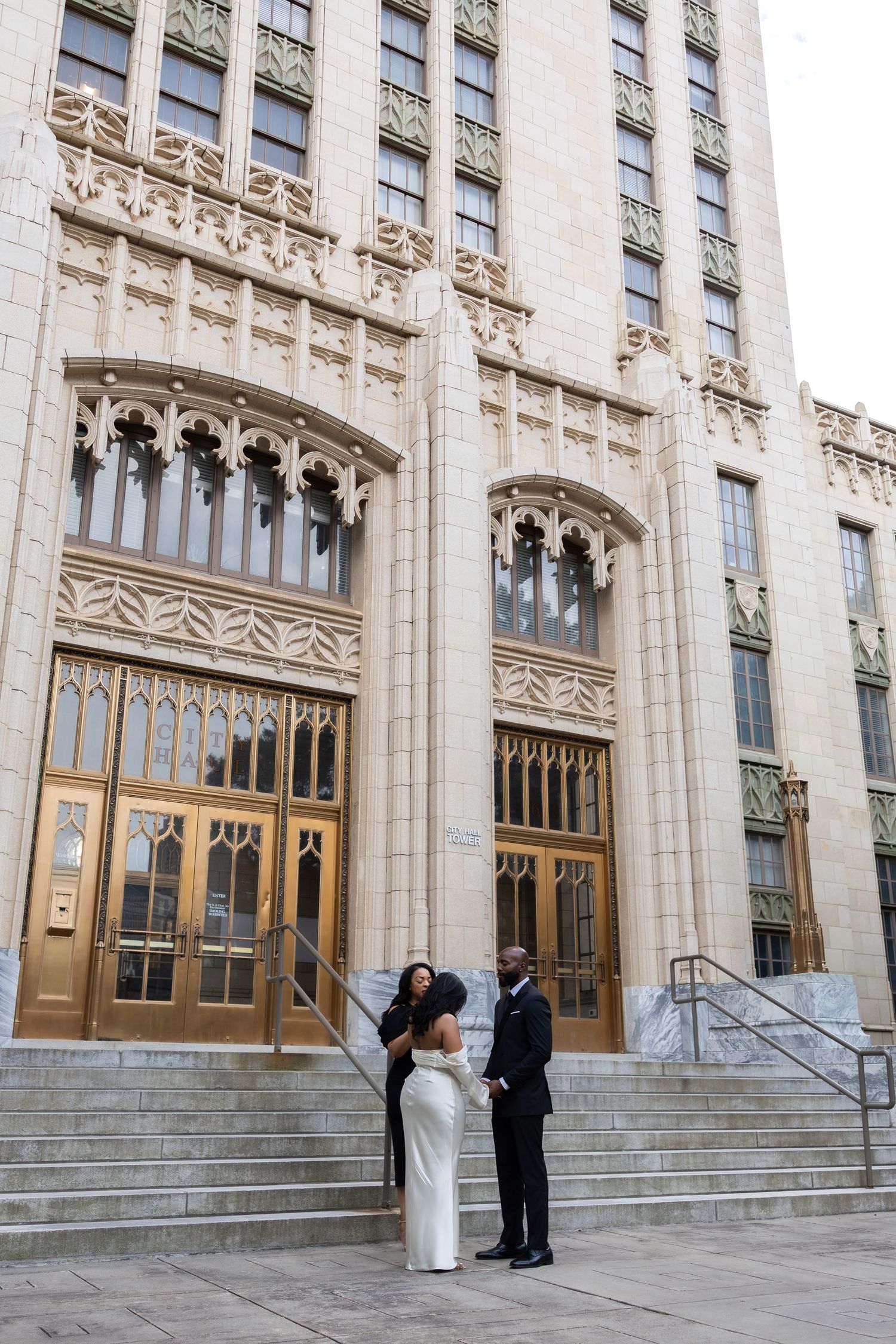 A couple in formal attire stands on steps before an ornate Art Deco building with golden doors.