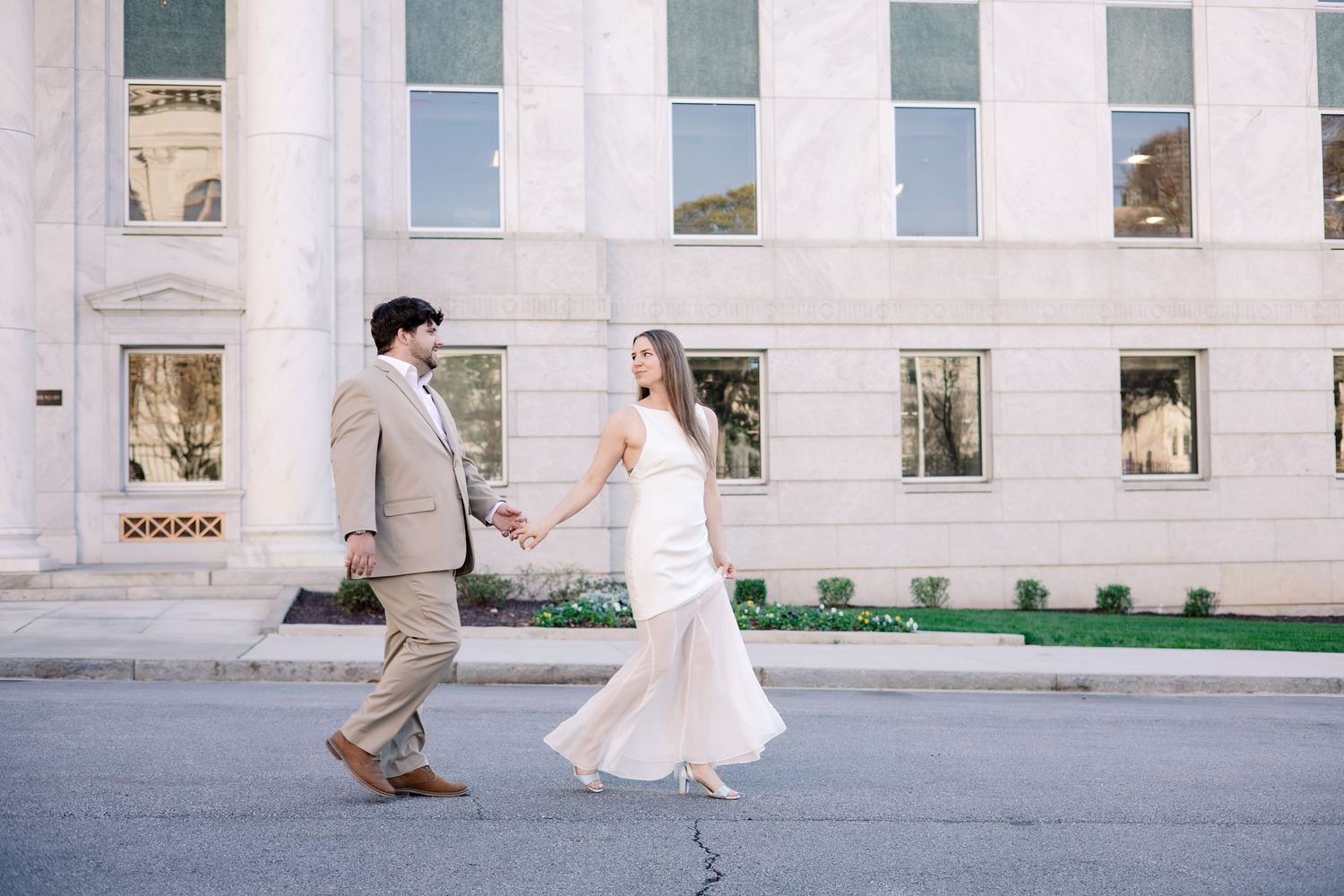 A couple in wedding attire playfully runs and dances along a white columned building exterior.