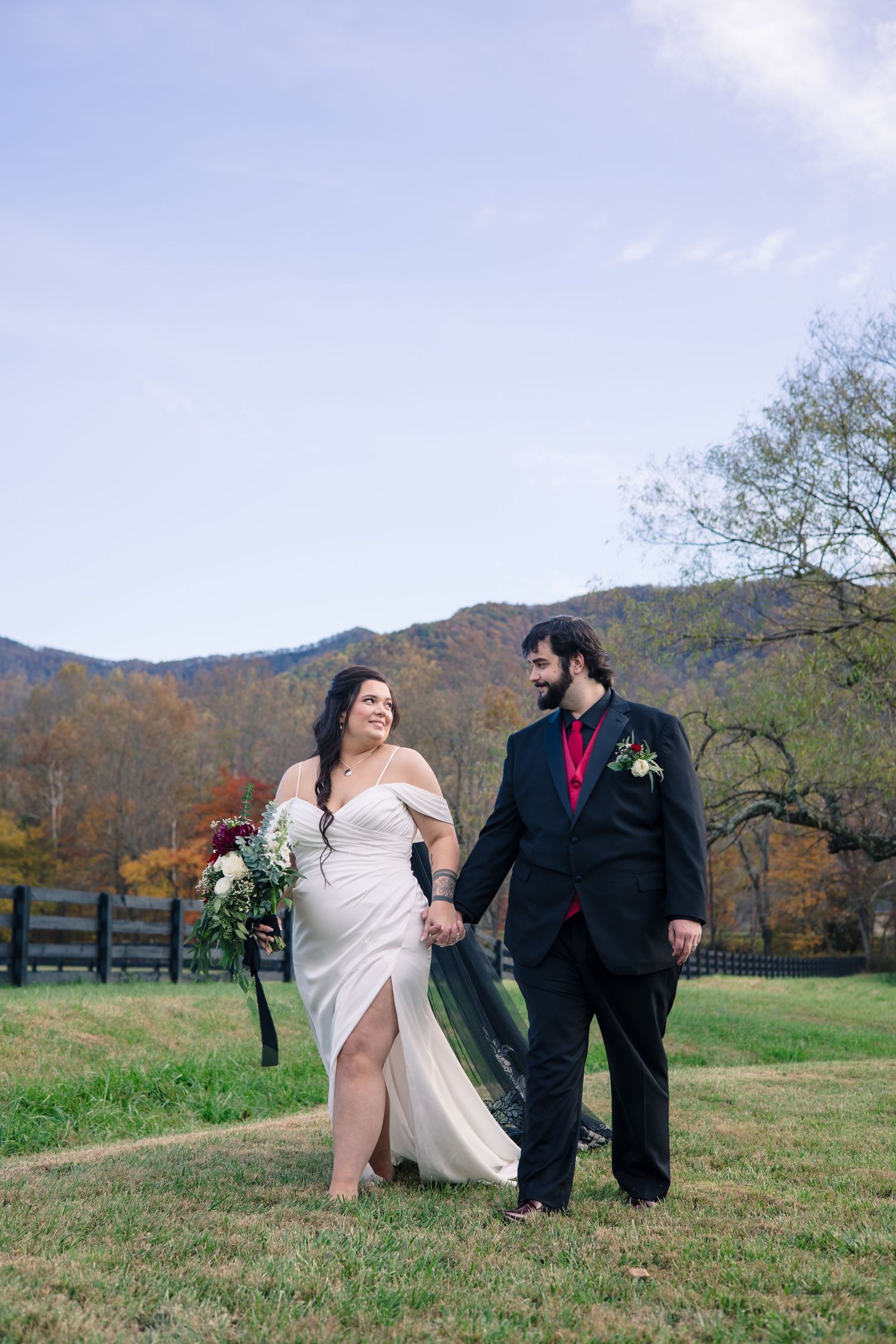 Newlyweds walking hand in hand across a grassy field with mountains in the background.