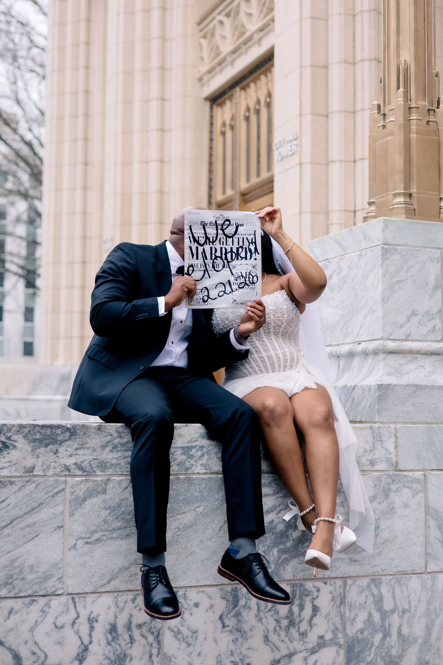 A couple sits on marble steps holding a sign while dressed in formal wedding attire.