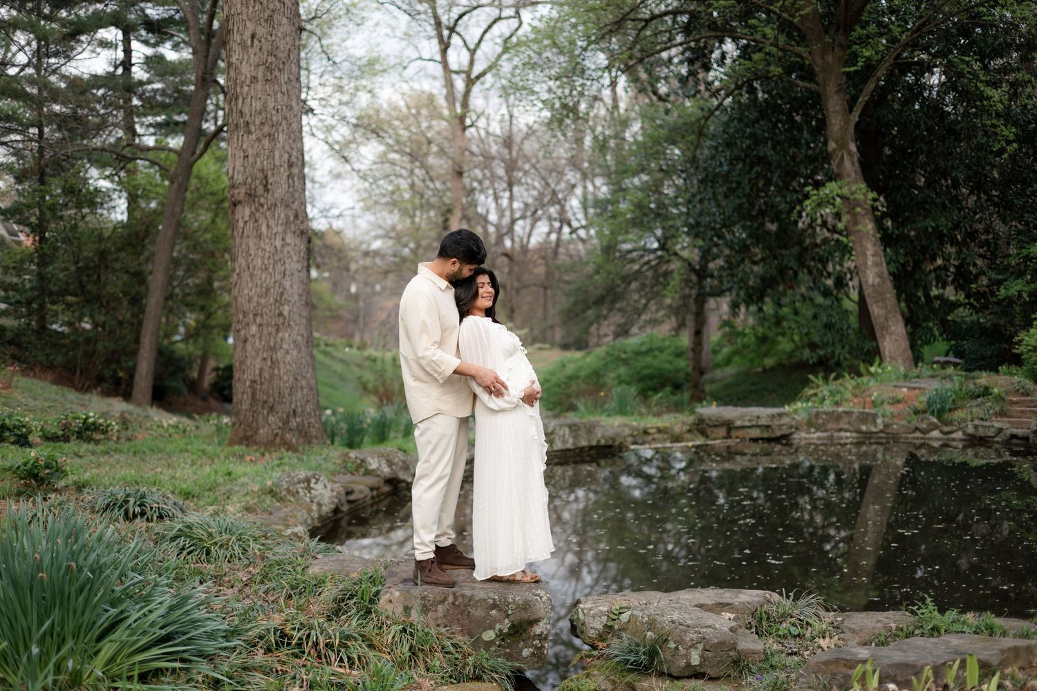 A romantic couple in white outfits embraces in a serene garden setting surrounded by tall trees and spring foliage.