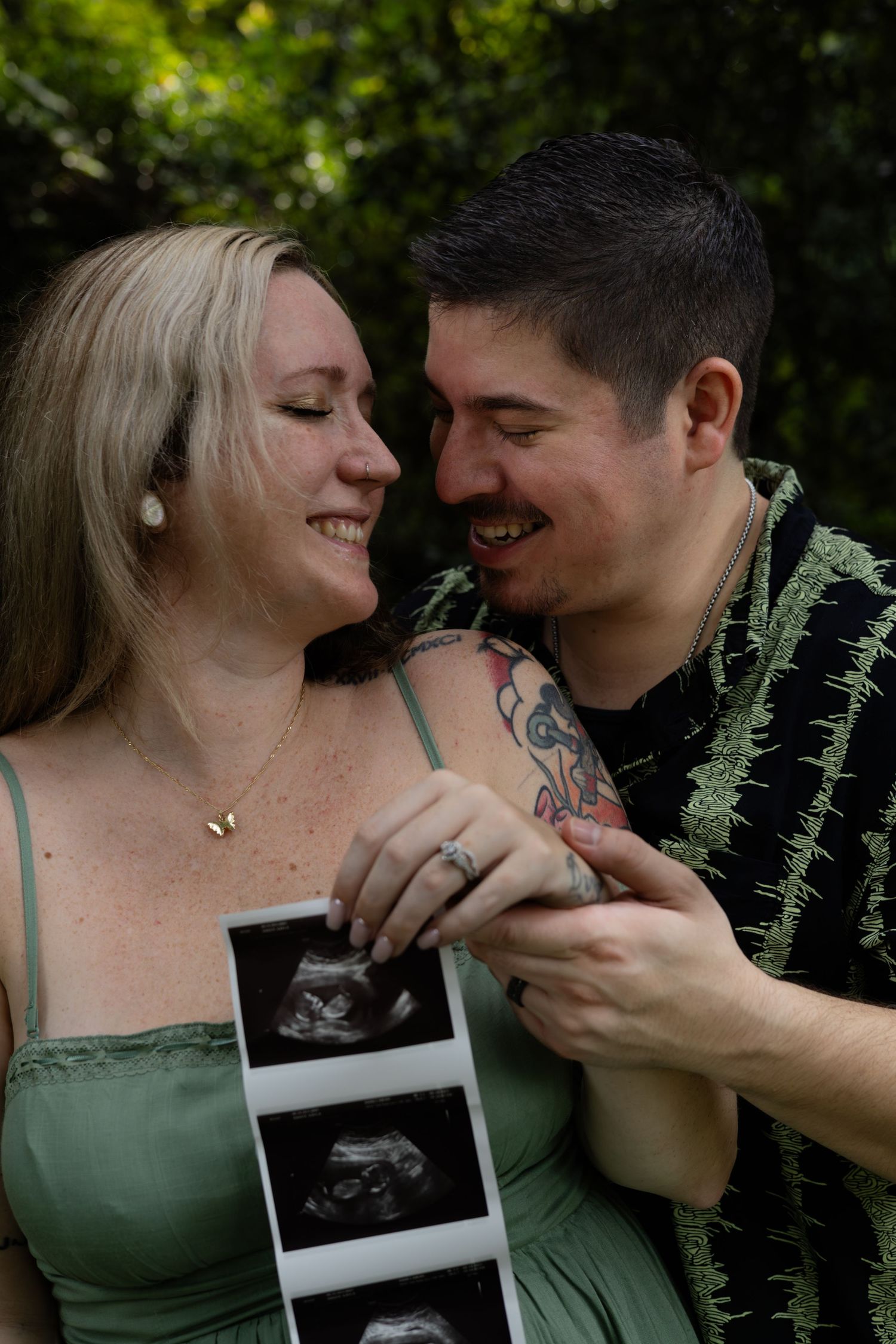 Couple shares intimate moments outdoors while holding ultrasound photo in green natural setting.