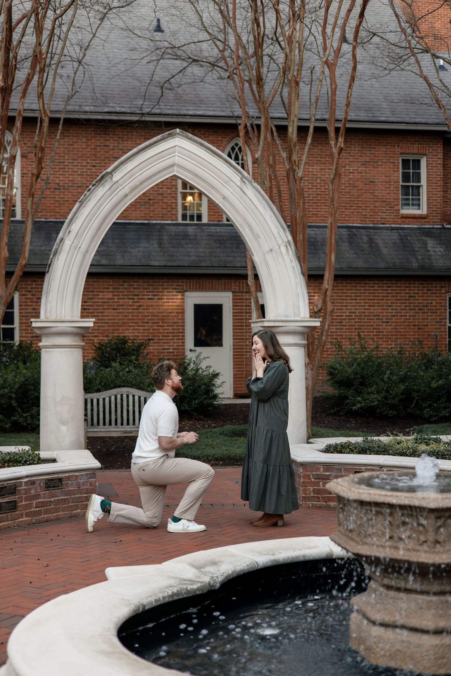 A proposal moment captured by a brick building with white archways and fountain.