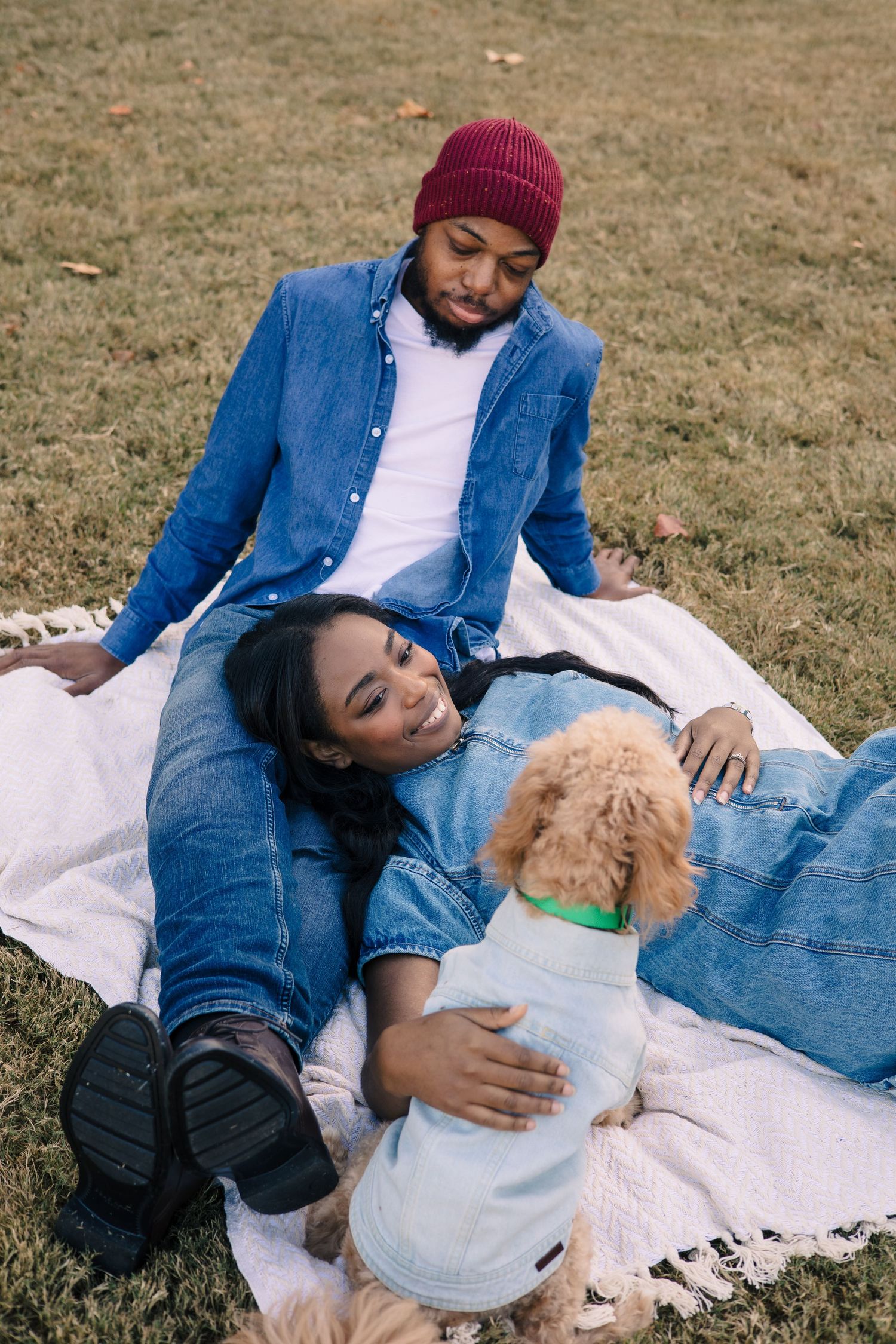 A couple relaxes on a white blanket in the grass with a small dog.