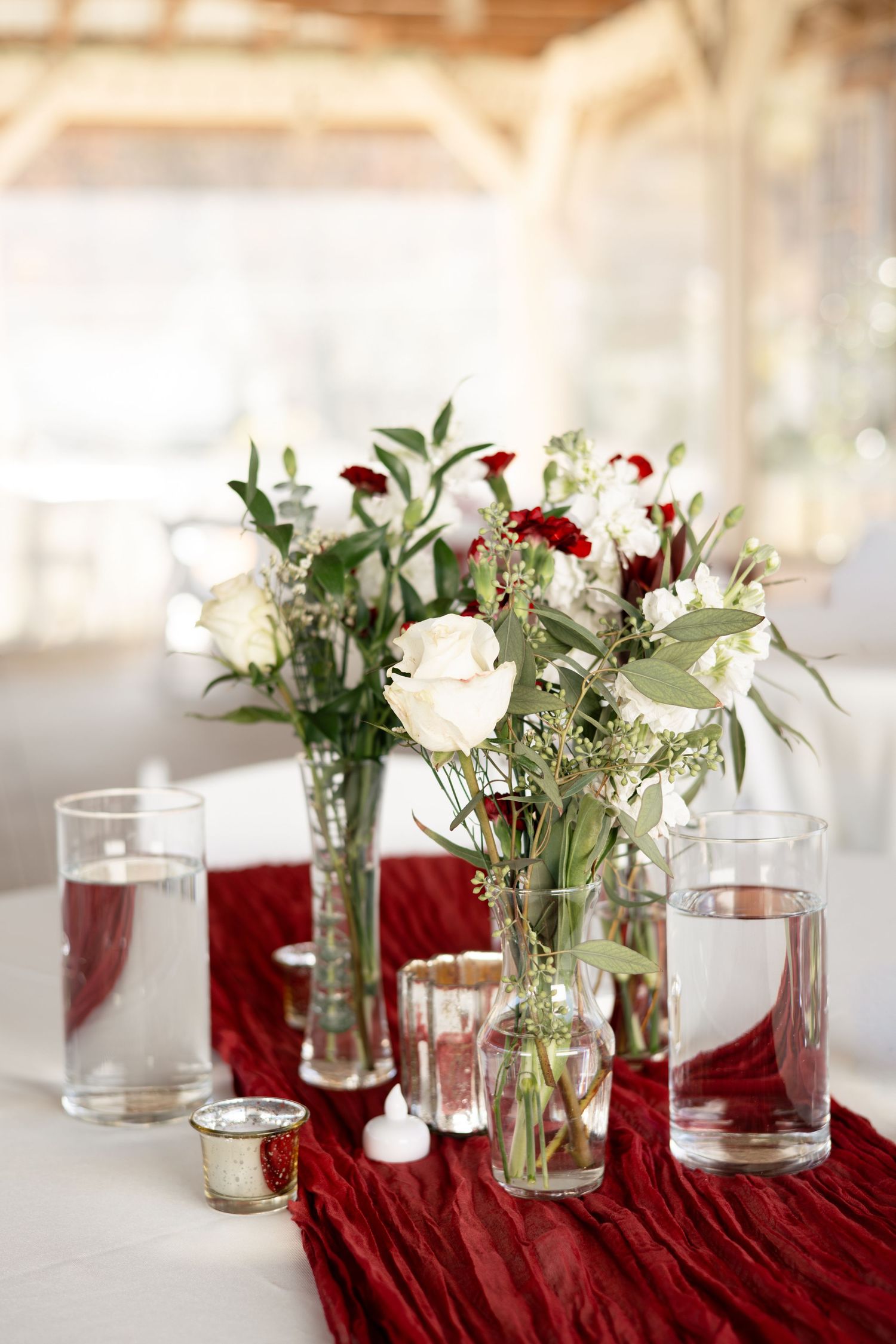 Wedding table decor with red velvet runner and glass vases holding white flowers and greenery.