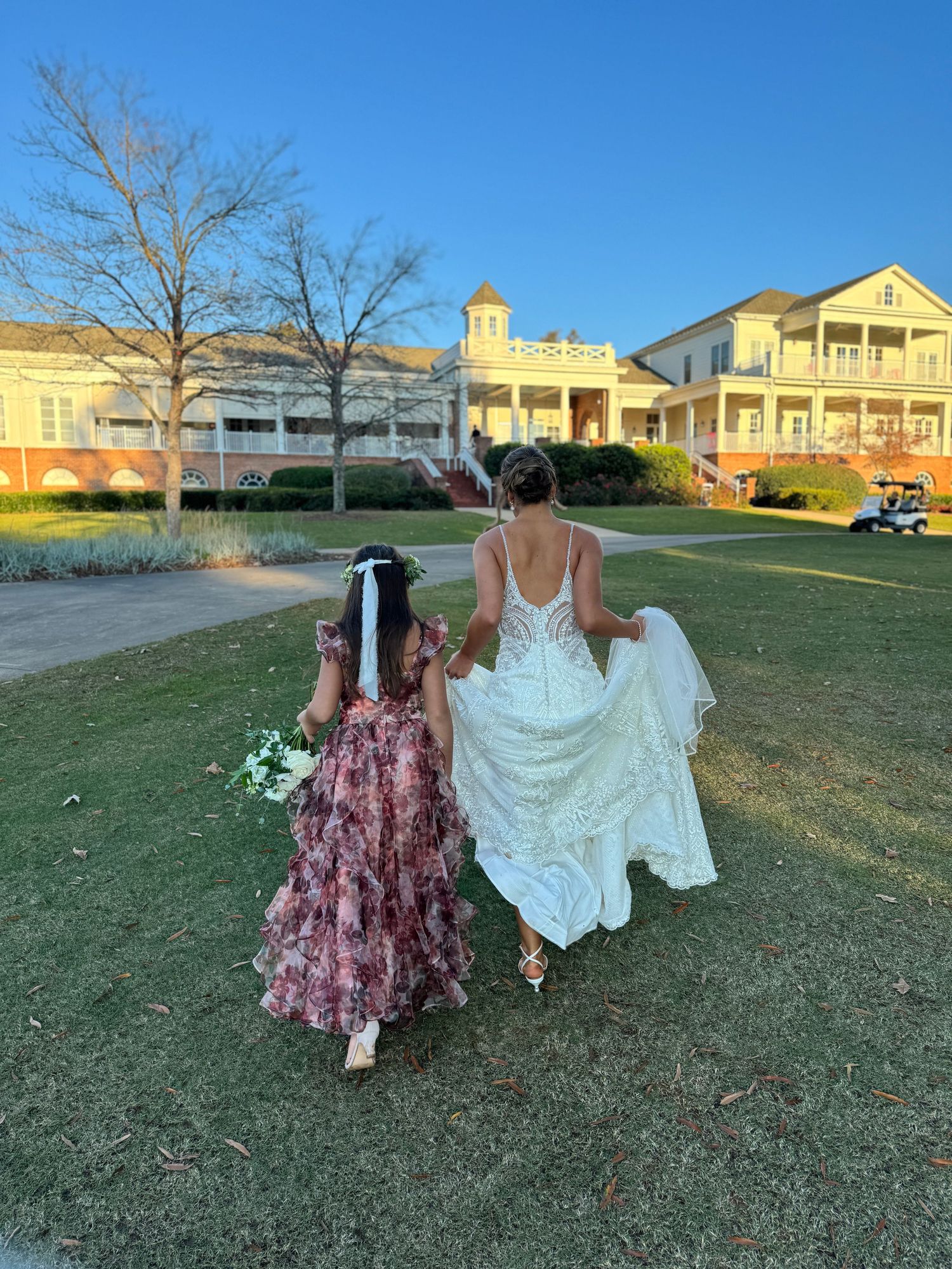 Wedding party members walk across a grassy field at sunset with buildings in the background.