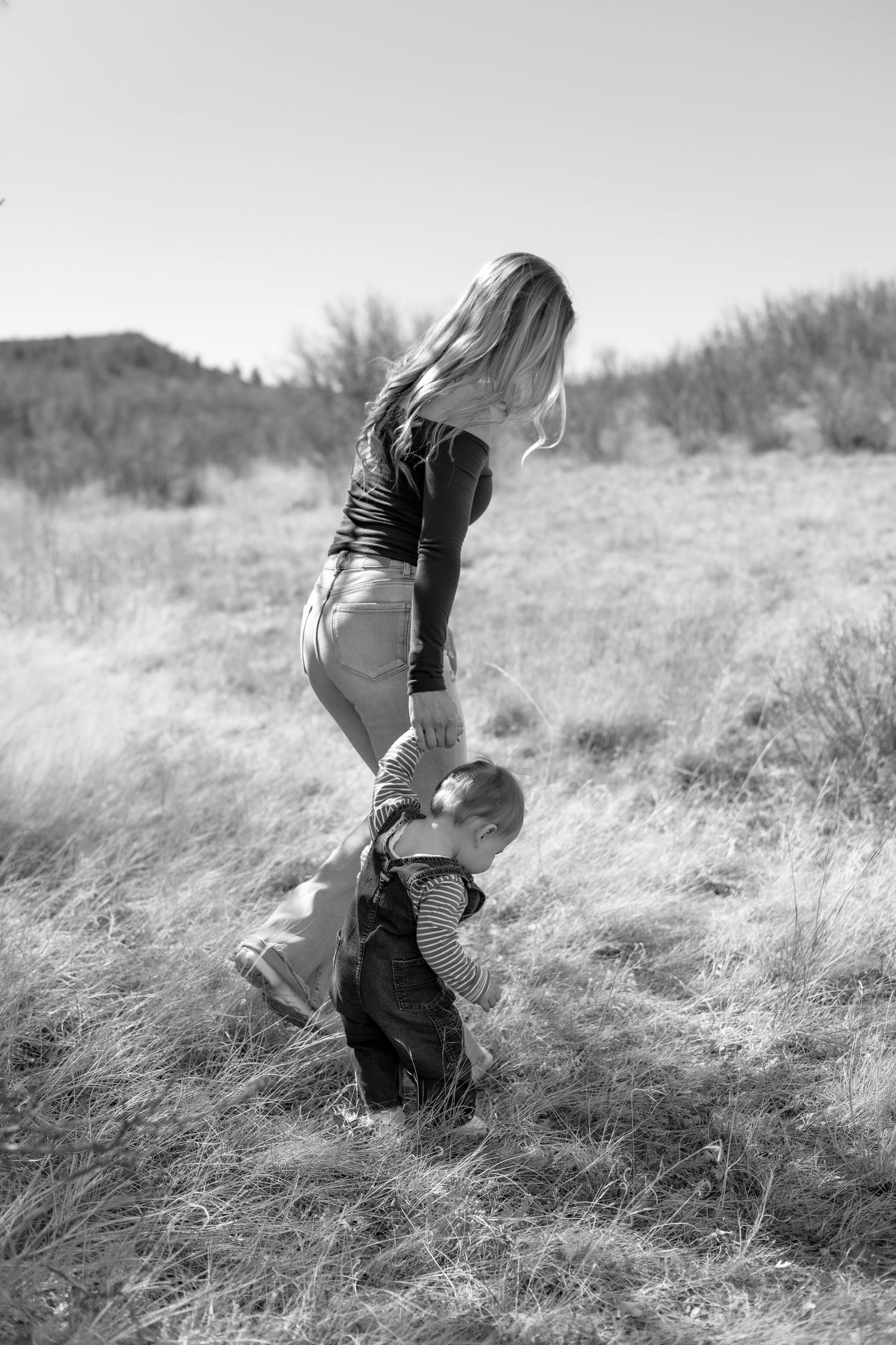 Black and white photo of a mother helping her toddler walk through a grassy field on a sunny day.
