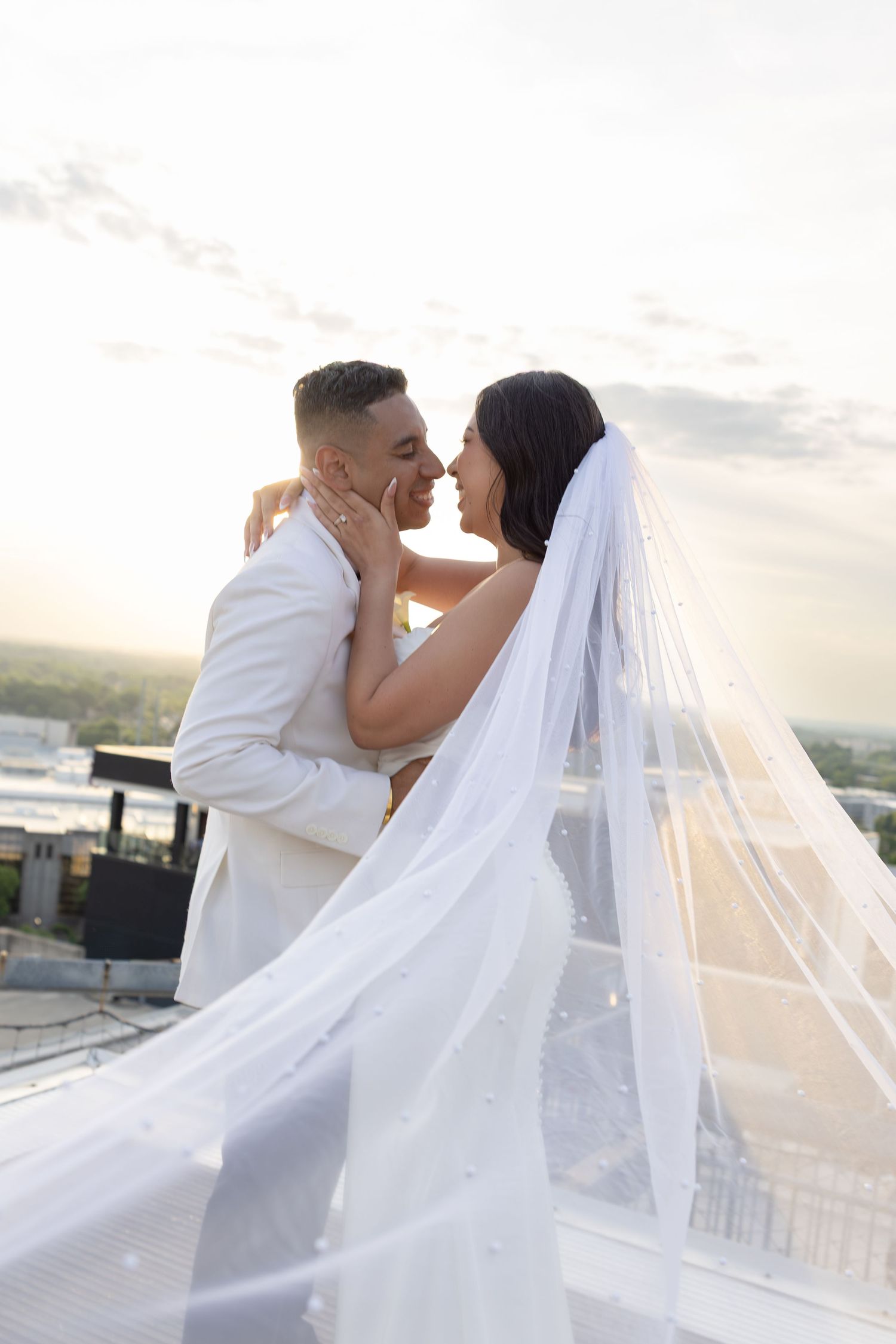 Bride and groom share a romantic almost-kiss on a rooftop, bride's long veil flowing in the breeze at sunset.