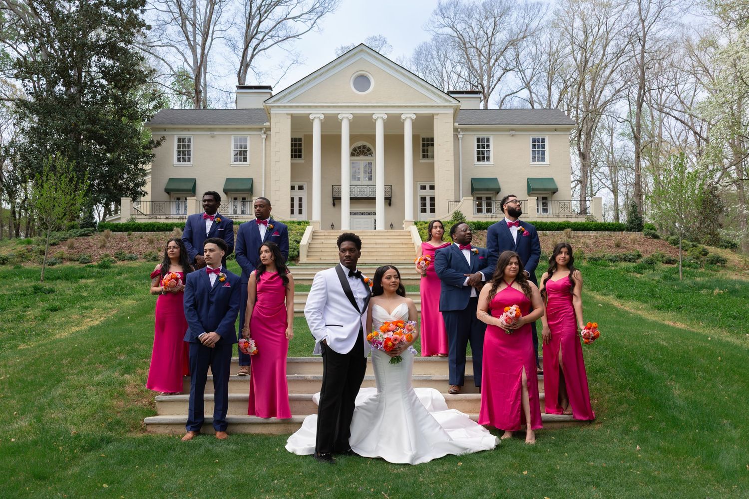 Wedding party poses on mansion steps, bridesmaids in pink gowns, groomsmen in navy suits, bride and groom centered.