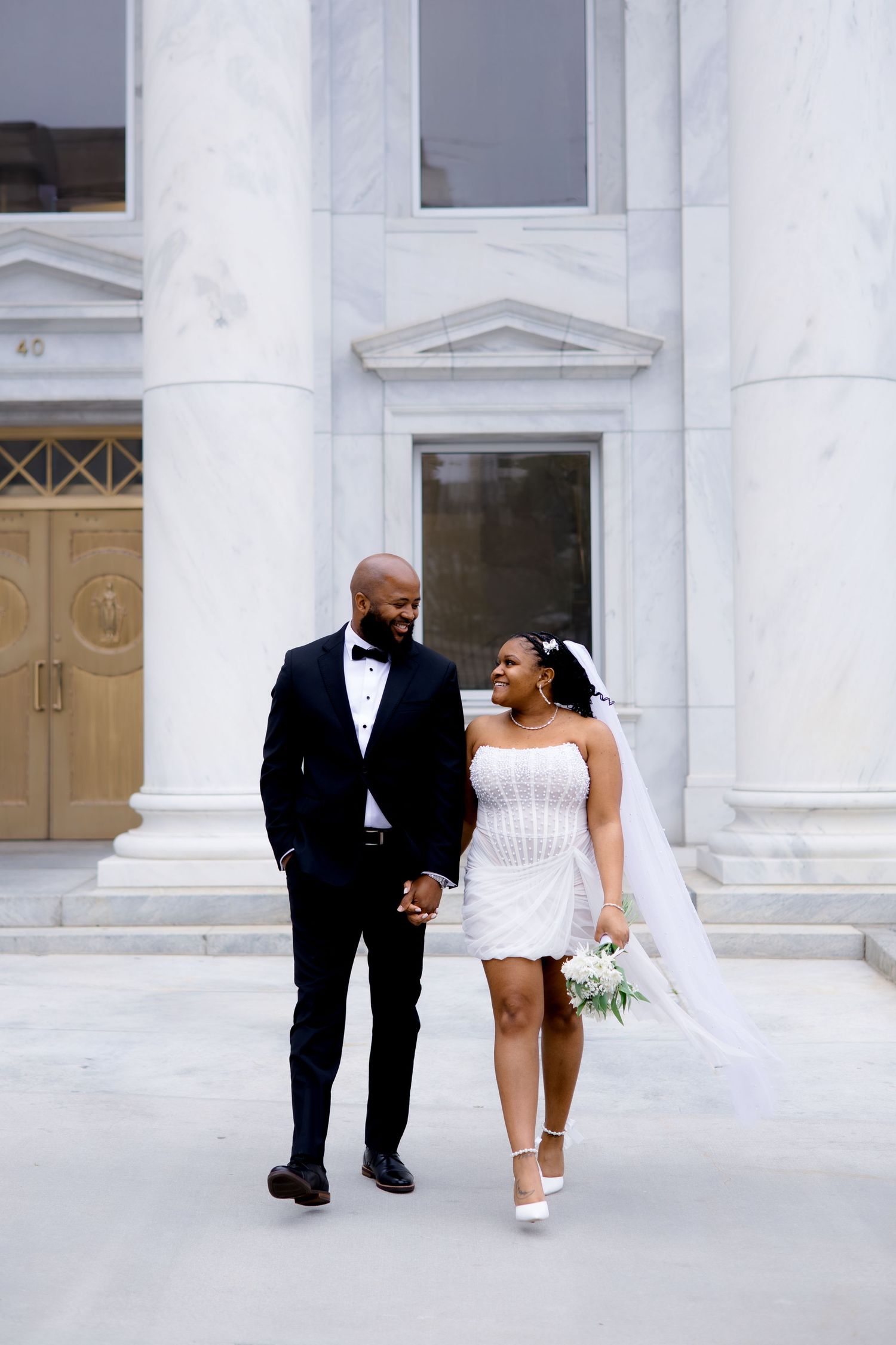A bride and groom in formal attire stand in front of an ornate building, holding hands and smiling at the camera.