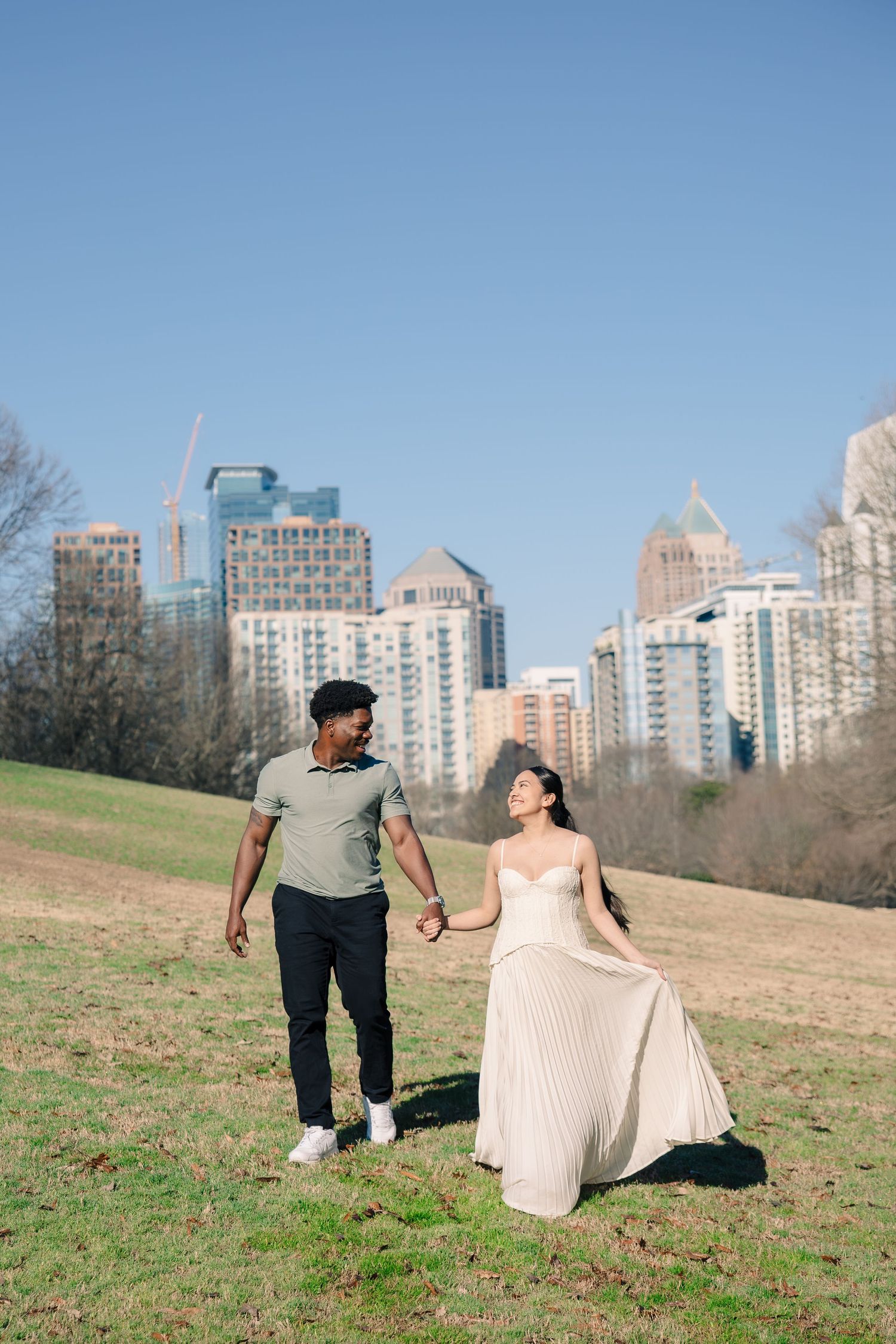 A couple in formal attire walks and dances together on a grassy hillside with city buildings visible in the background.