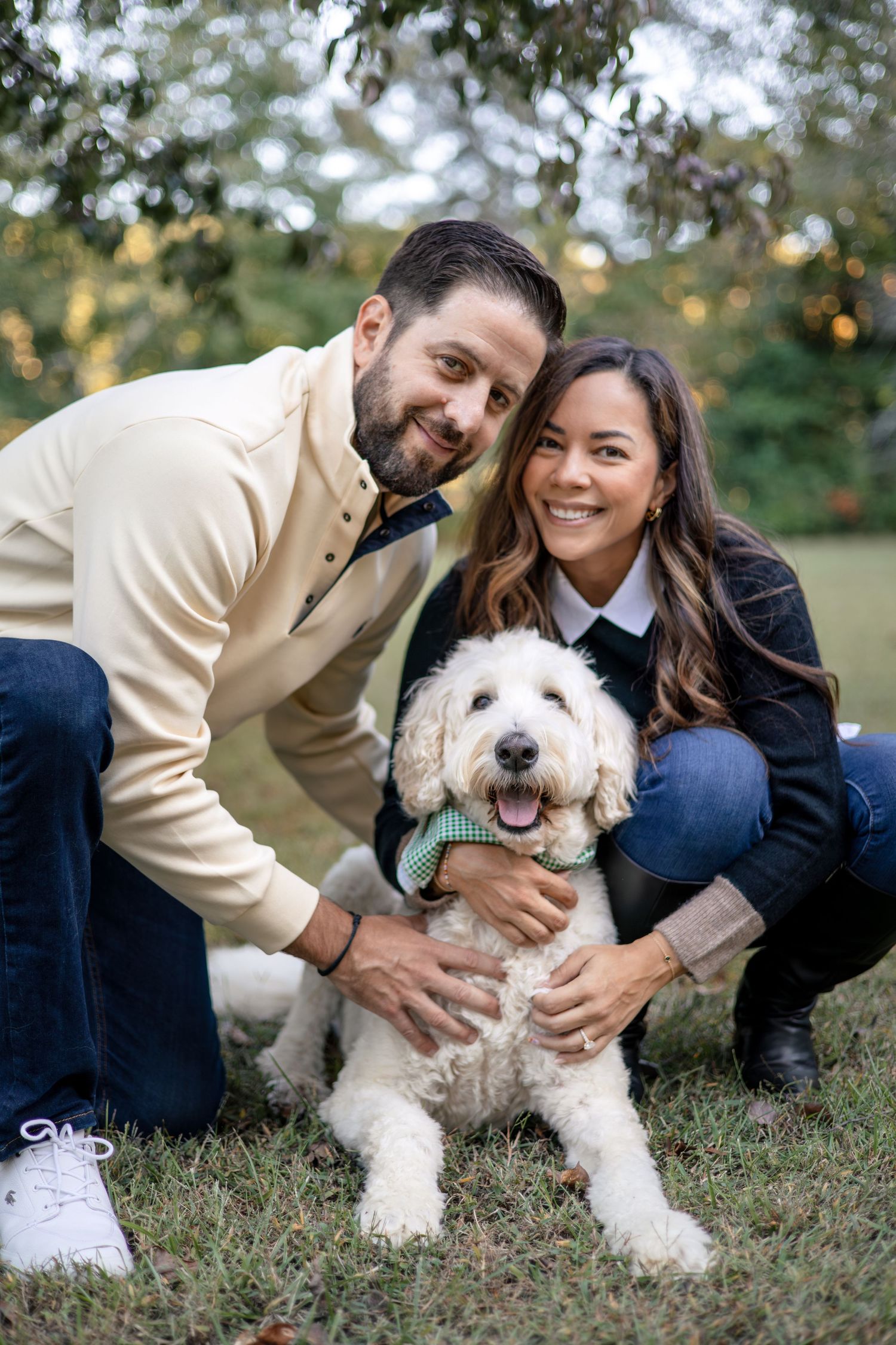A happy couple poses outdoors with their fluffy white dog on a sunny day.