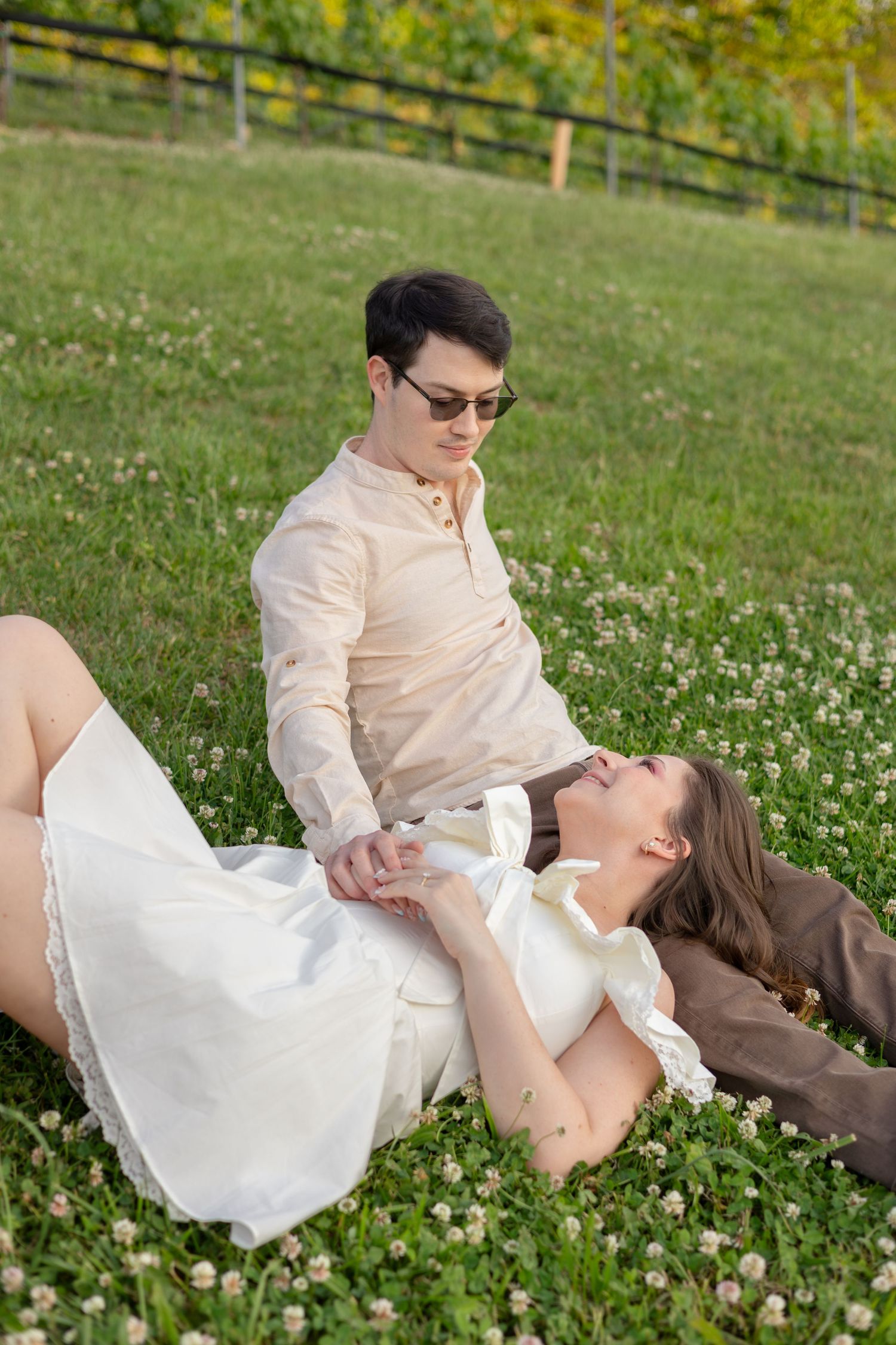 A couple relaxing on green grass, woman in white dress lying down holding hands with man in beige shirt wearing sunglasses.