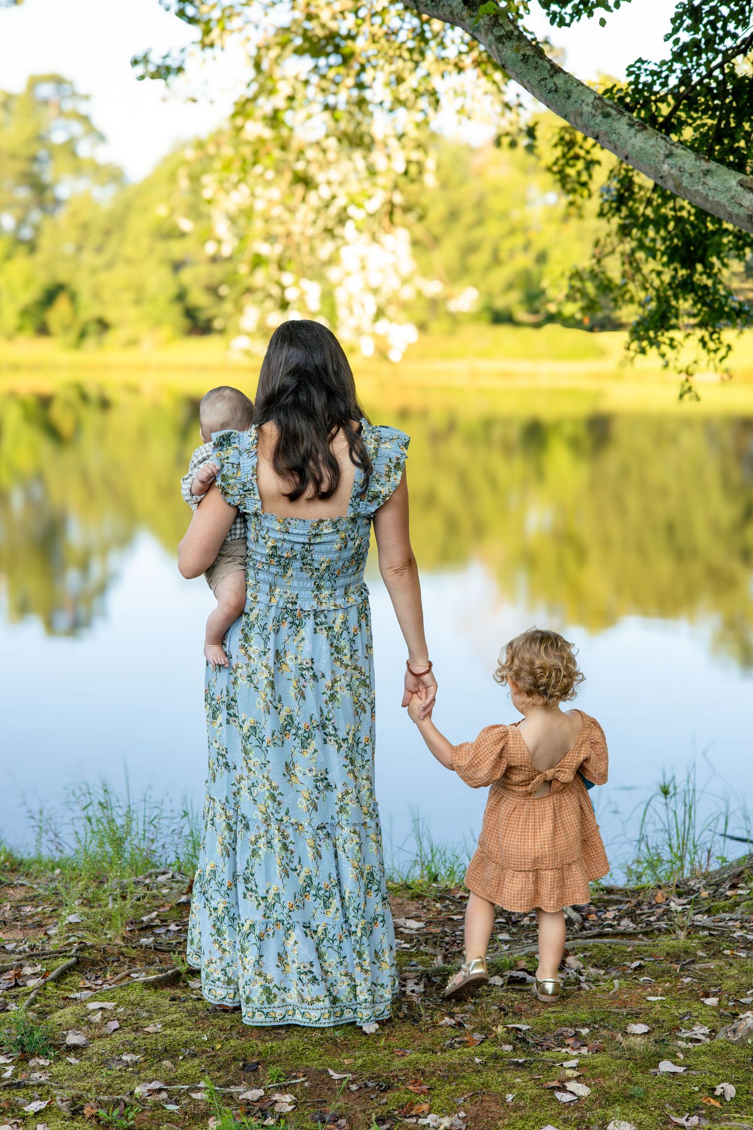 Mother and child sharing tender moments by a tranquil lakeside under green trees.