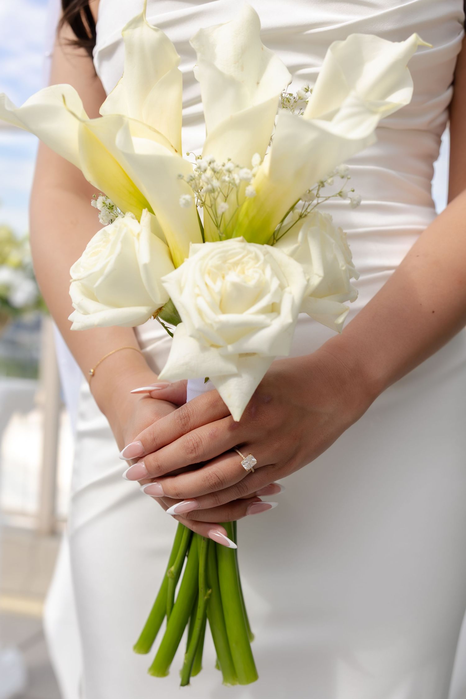 Bride holding elegant white calla lily and rose wedding bouquet with baby's breath, wearing diamond engagement ring.
