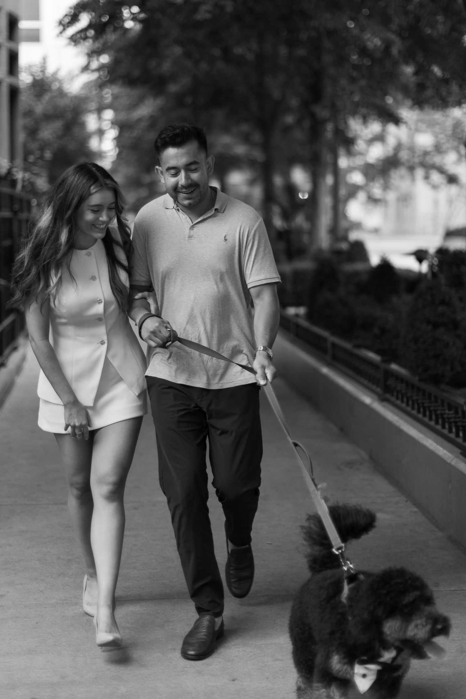 A couple walking their small fluffy black dog on a paved pathway in the park, captured in black and white.